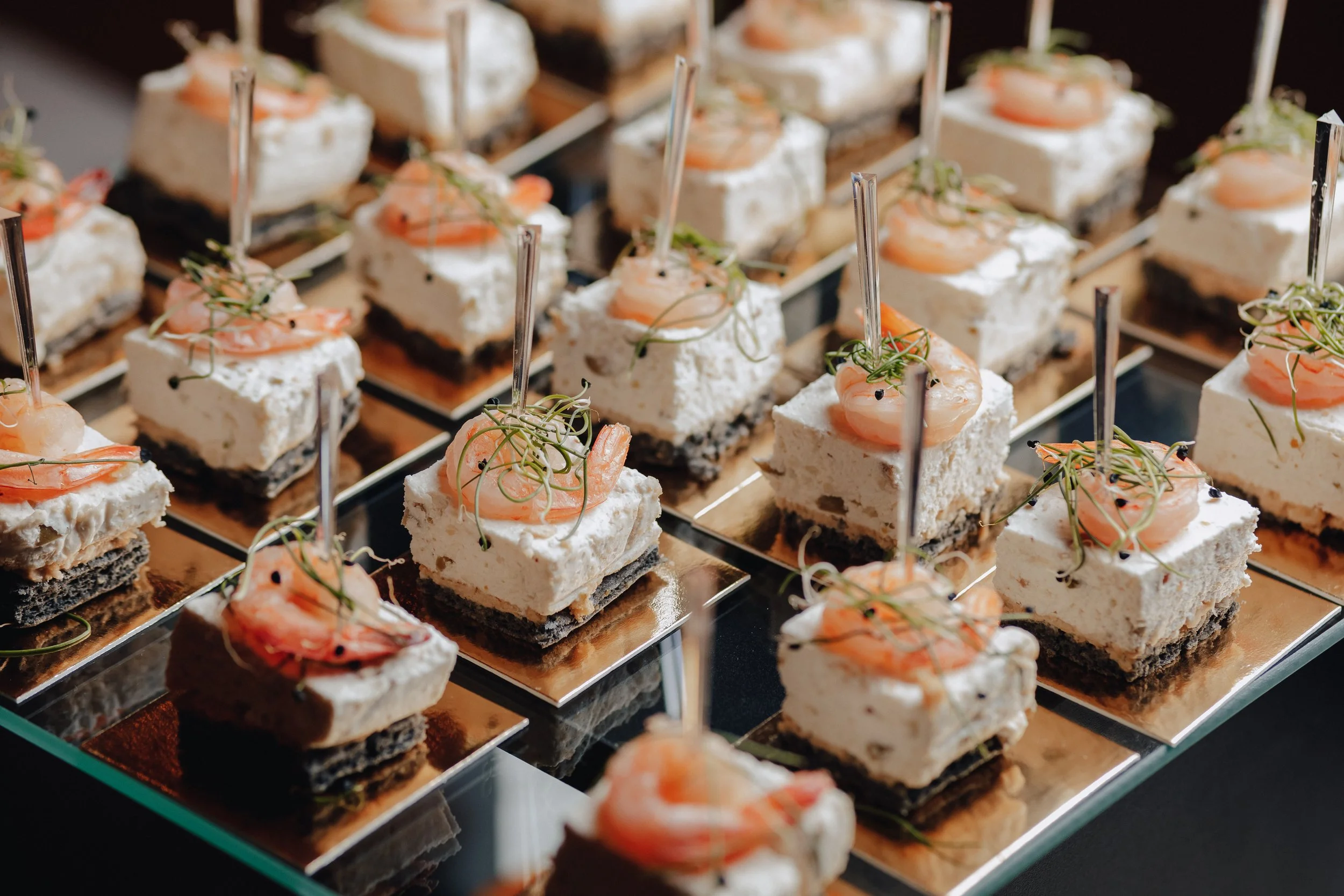 Small square desserts with layers of white cream, topped with shrimp and green microgreens, arranged on a reflective surface.