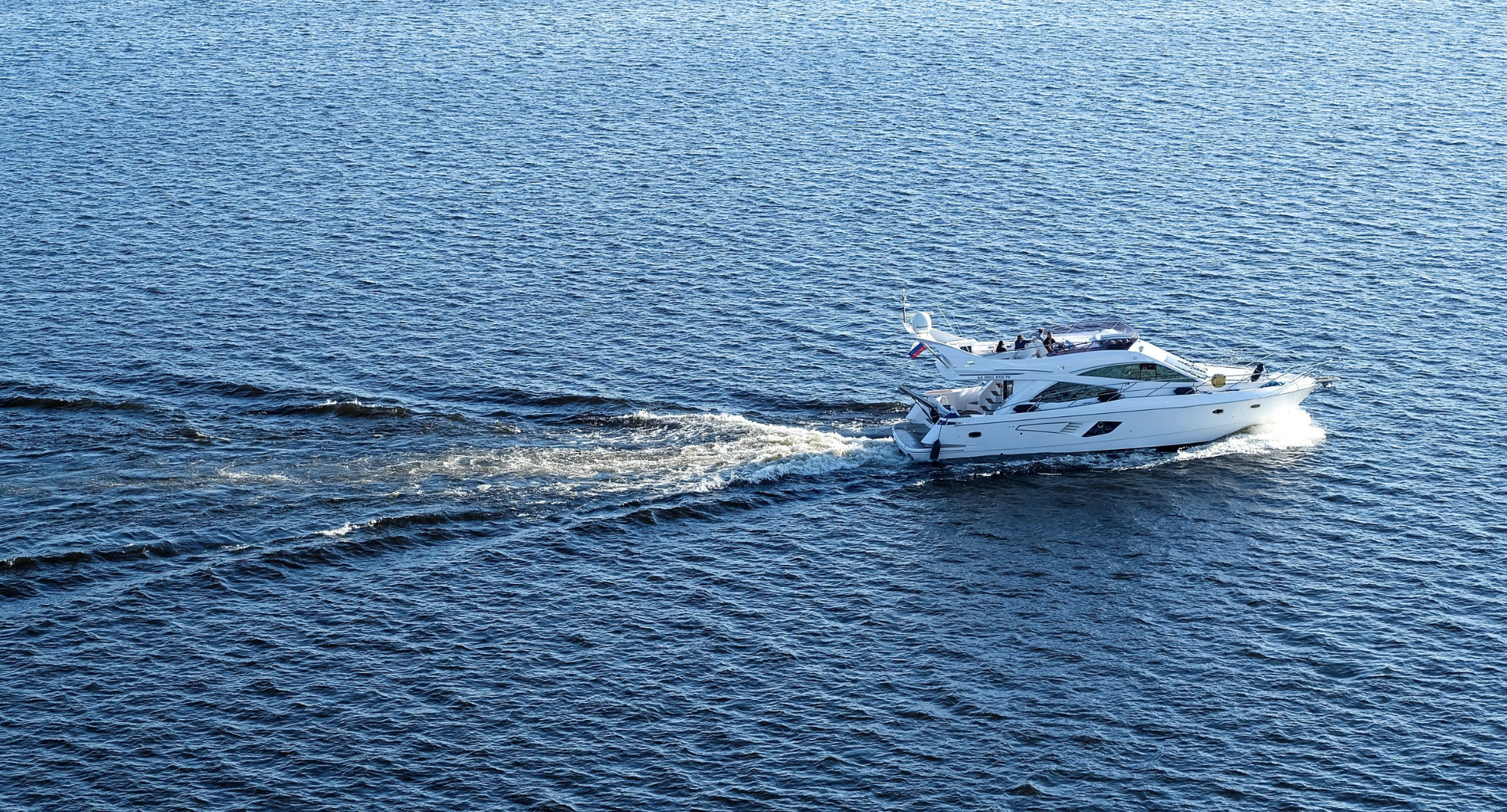 A white motor yacht cruising on calm blue ocean water with a few people on the deck.
