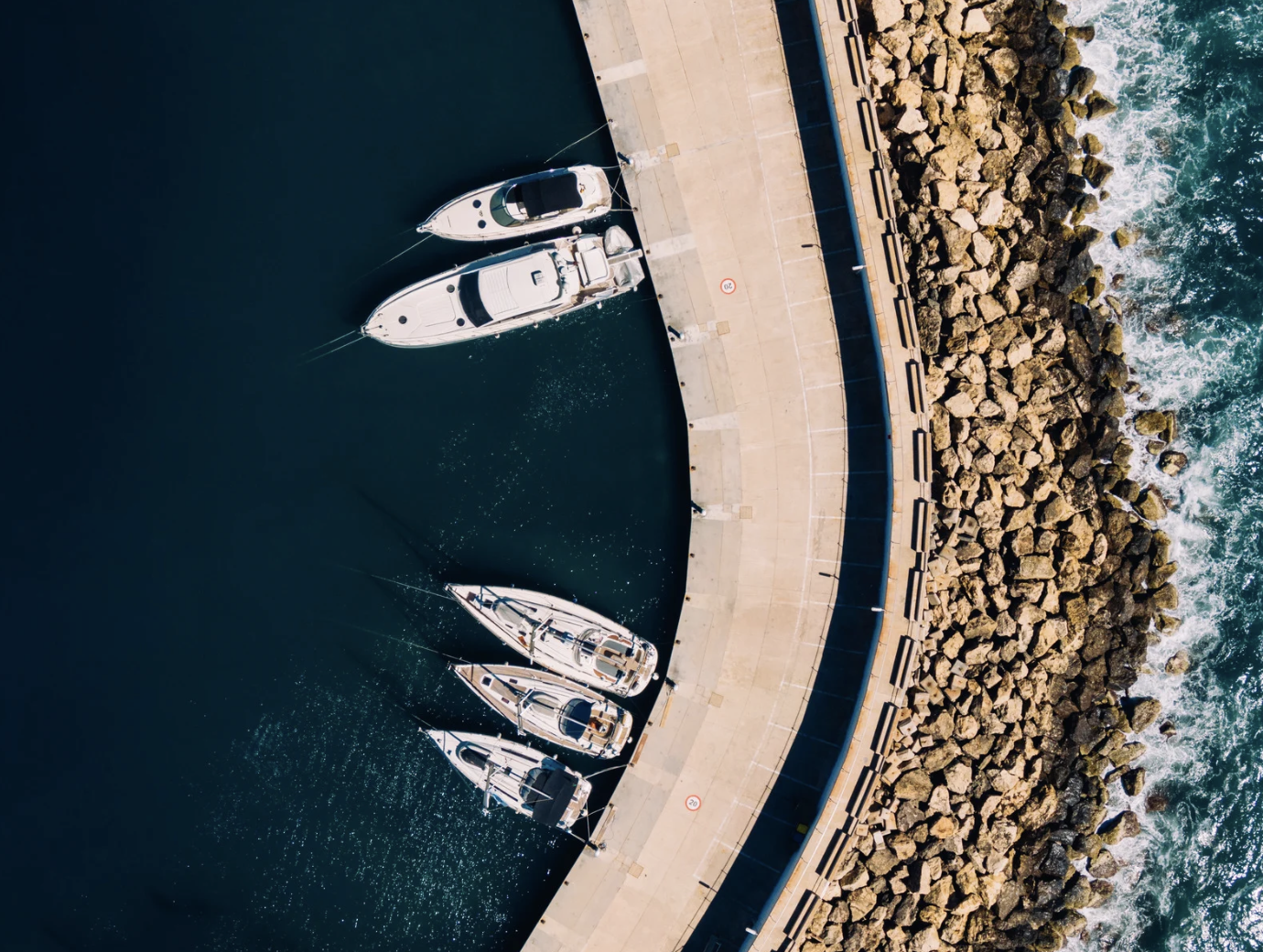 Aerial view of a pier with five white yachts docked, rocks along the shoreline, and ocean waves crashing against the rocks.