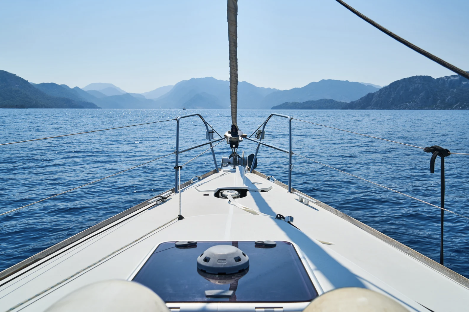 From the bow of a sailboat looking out on calm blue water and distant mountain ranges under a clear sky.