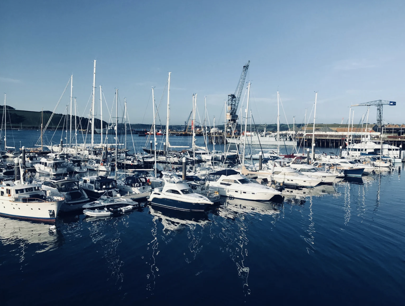 A marina filled with various boats and yachts docked on calm water, with cranes and industrial structures in the background under a clear blue sky.