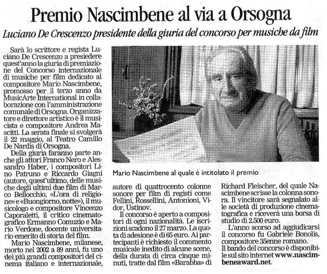 Fotografia di un uomo anziano con capelli bianchi, seduto a un tavolo, che sorride e guarda verso l'obiettivo, con una tazza e alcuni documenti di fronte.