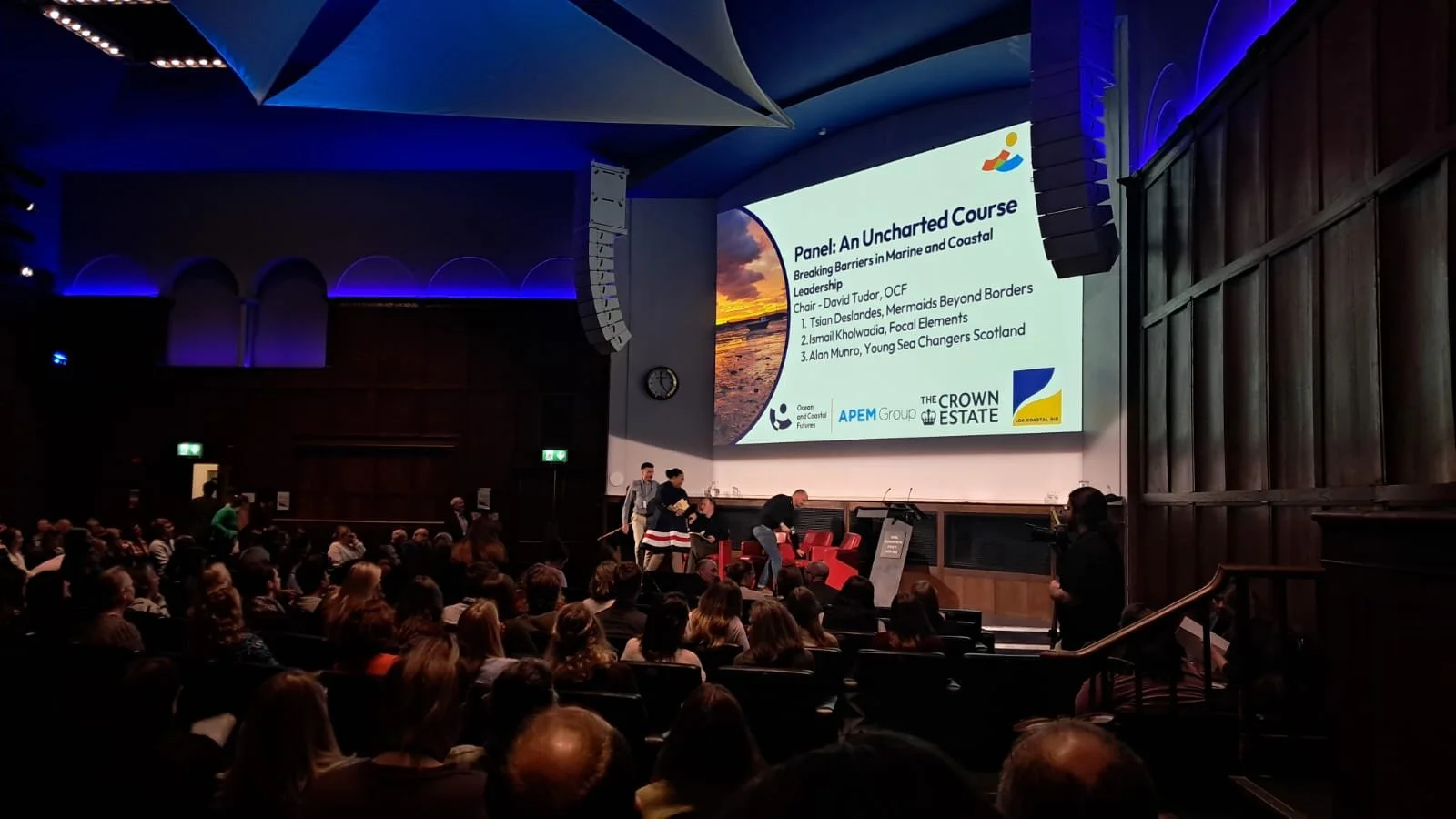 A conference or panel event in a large auditorium with a speaker preparing on stage. A large screen displays the presentation titled "Panel: An Uncharted Course" about breaking barriers in marine and coastal leadership.