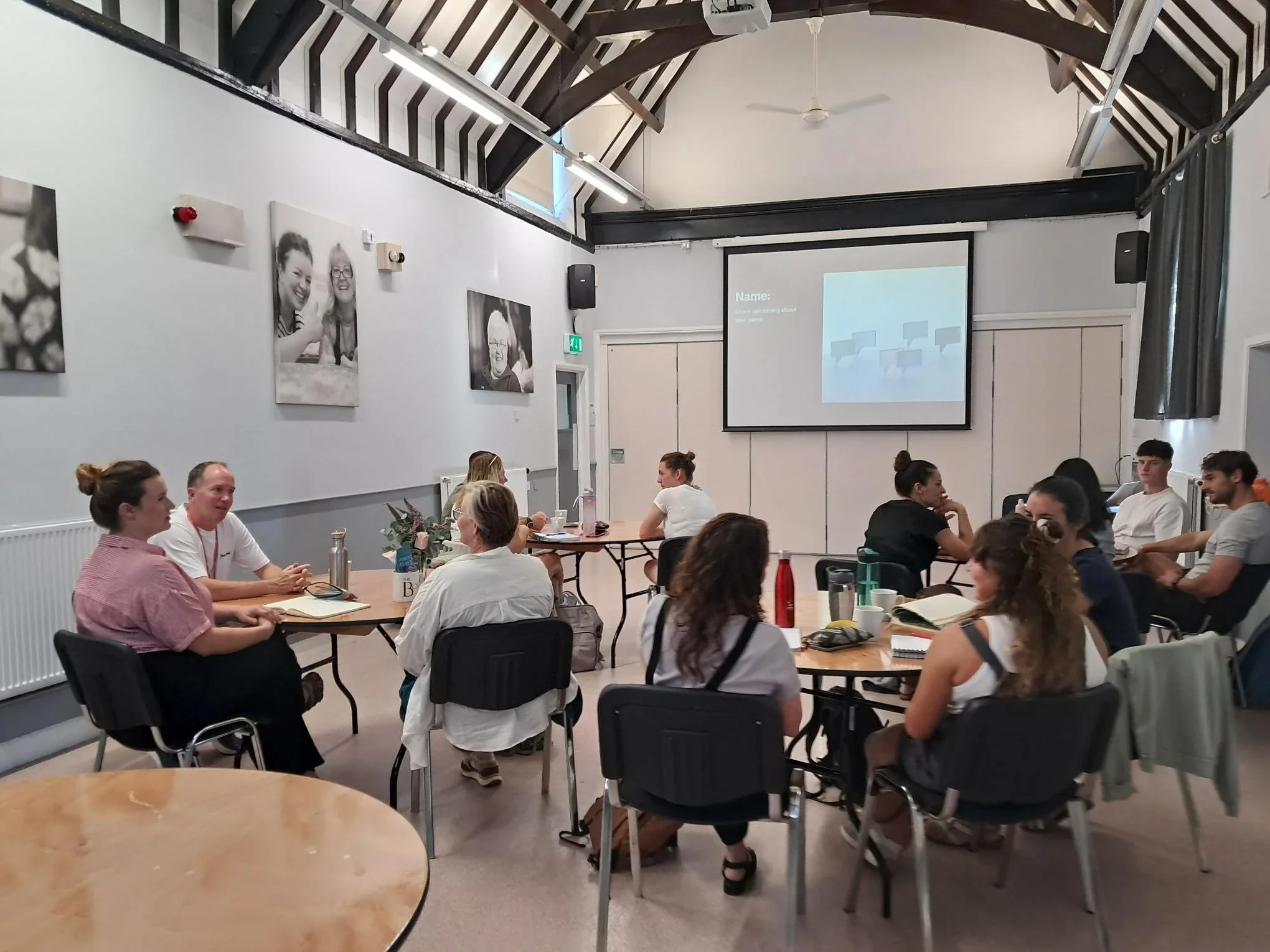 A group of people seated around tables in a meeting room, engaged in discussion, with a projector screen displaying a presentation at the front.