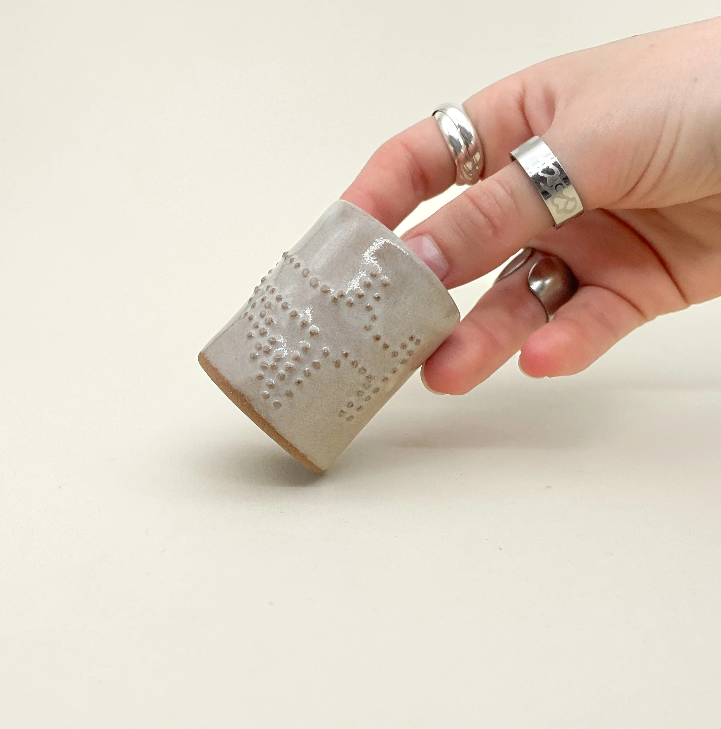 A hand holding a beige ceramic mug with embossed dotted patterns, wearing silver rings, against a plain white background.