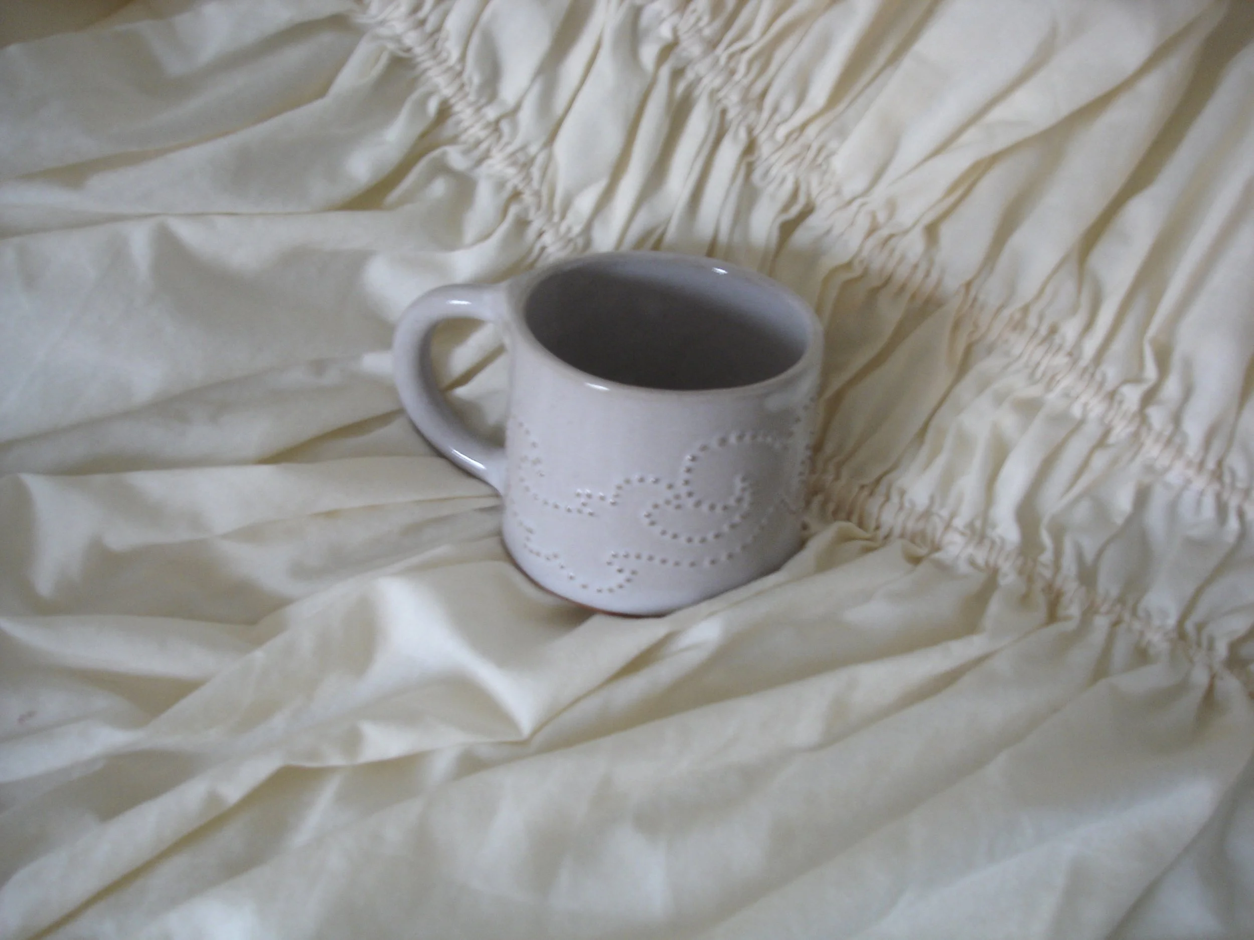 A white ceramic mug with embossed decorative swirls, sitting on a cream-colored, rumpled fabric surface.