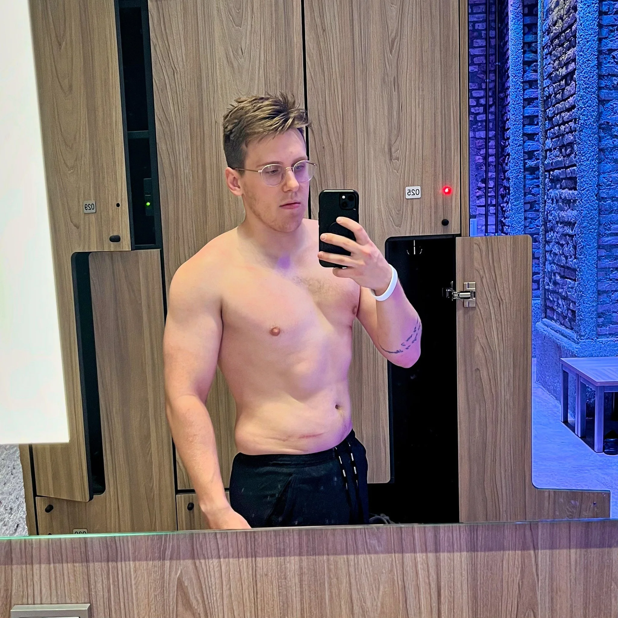 Zachary Rioux with glasses and light brown hair taking a mirror selfie in a locker room. He is shirtless, wearing black shorts, and has a white wristband on his left wrist.