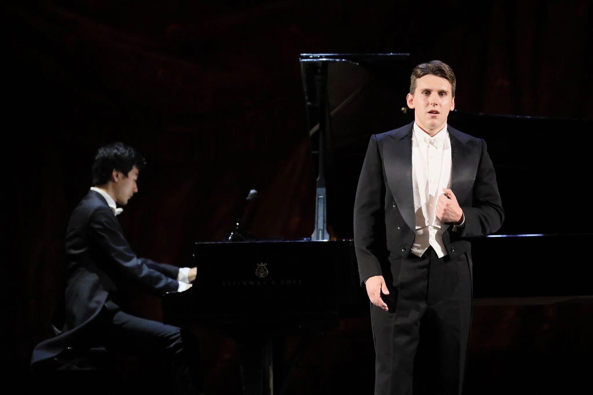 Opera singer, Zachary Rioux, in a tuxedo performs on stage with a pianist playing a grand piano in the background.