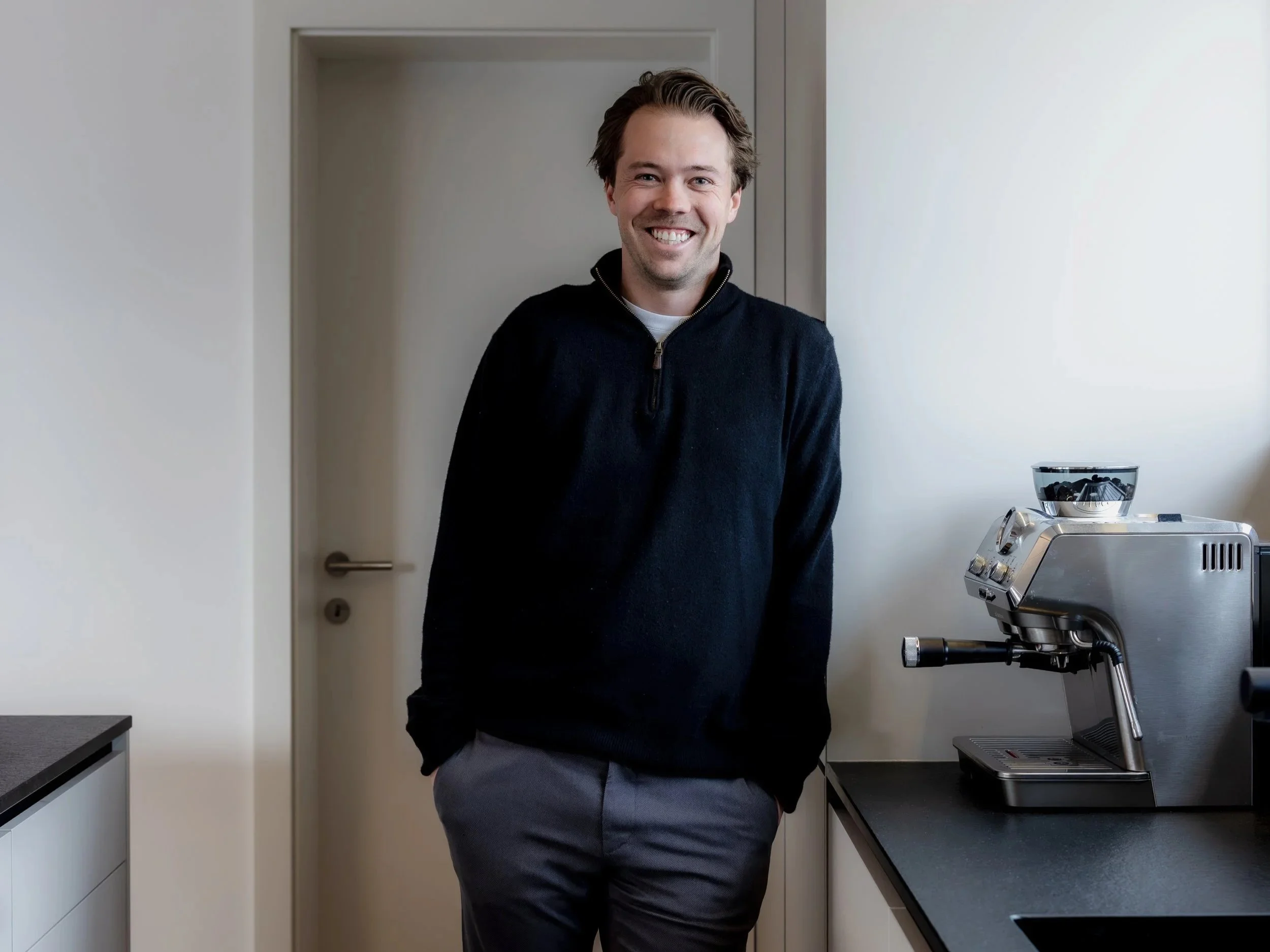 Ein Mann steht in einer modernen Küche neben einer Espressomaschine und lächelt in die Kamera.