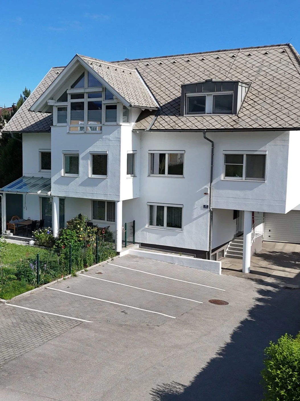Mehrstöckiges weißes Wohnhaus mit Balkon und Dachfenstern, Parkplatz im Vordergrund, blauer Himmel.