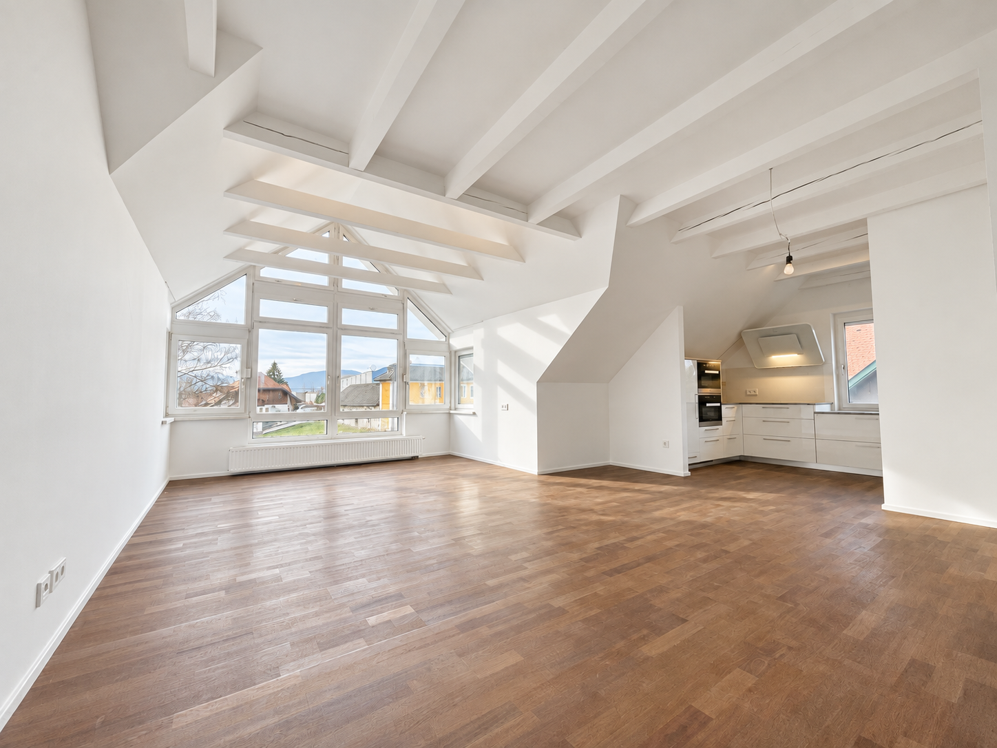 Ein großes, helles, und modernen Wohnzimmer mit Holzboden, weißen Wänden und großen Fenstern, die viel Tageslicht hereinlassen, mit offener Küchenbereich im Hintergrund.