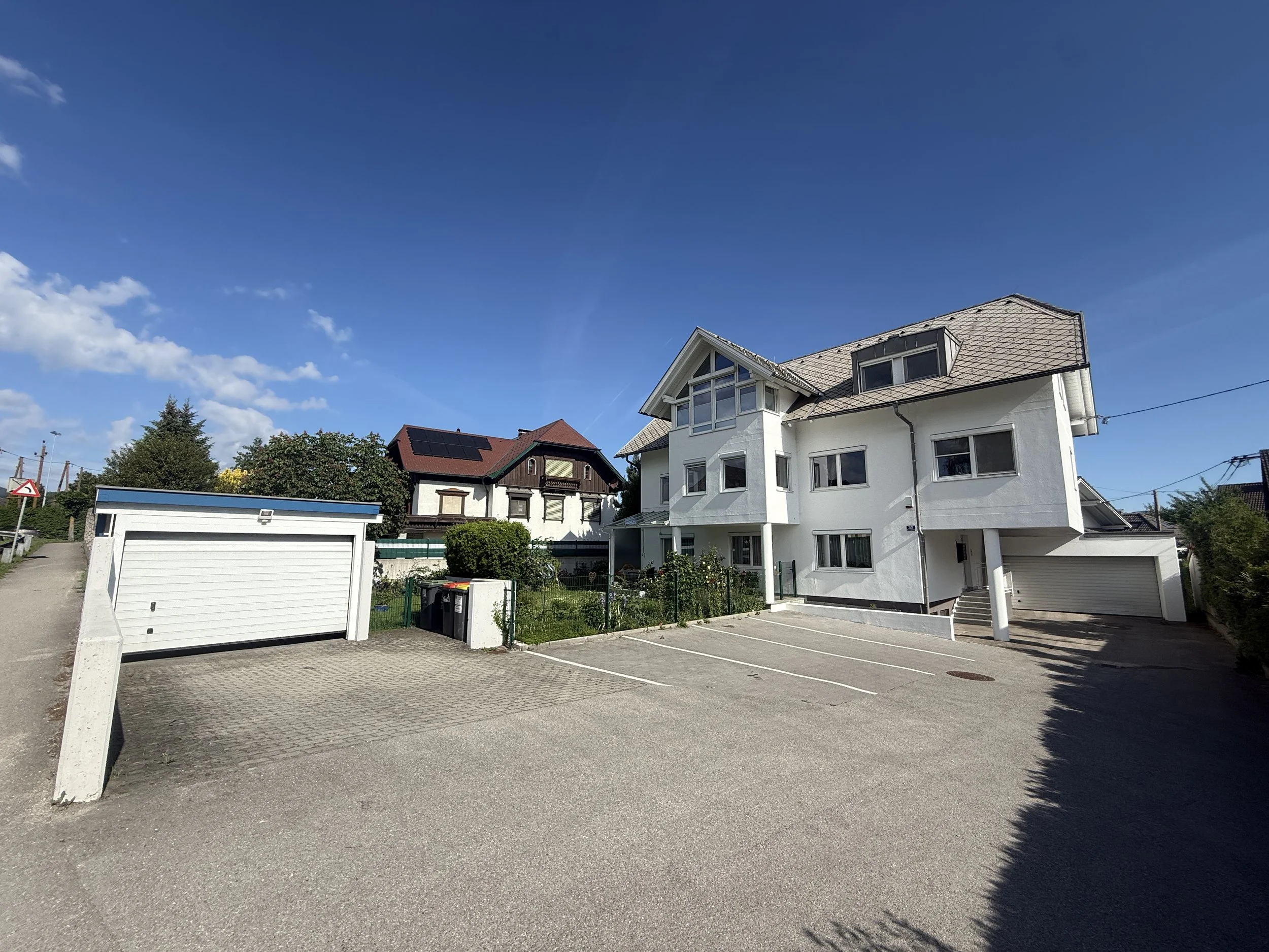 Ein Wohnhaus mit mehreren Stockwerken, weißen Wänden und großen Fenstern, darunter ein Carport. Der Himmel ist klar und blau mit wenigen Wolken, und es gibt Bäume im Hintergrund. Das Bild wurde an einem sonnigen Tag aufgenommen.