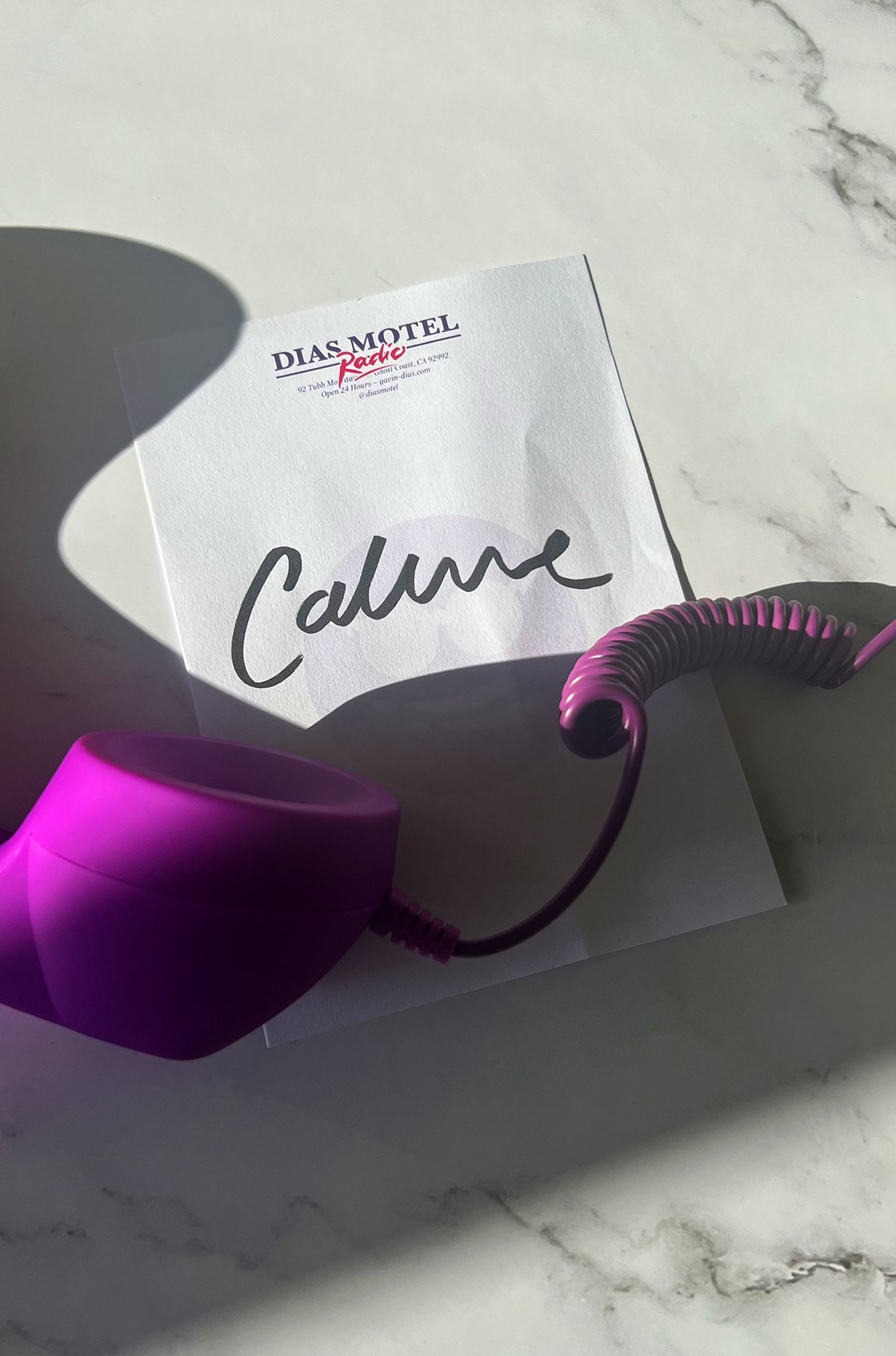 A white note paper on a marble surface, with the word 'Calm' written in black marker. The paper is casting a shadow, and there's a purple headphone on the surface.