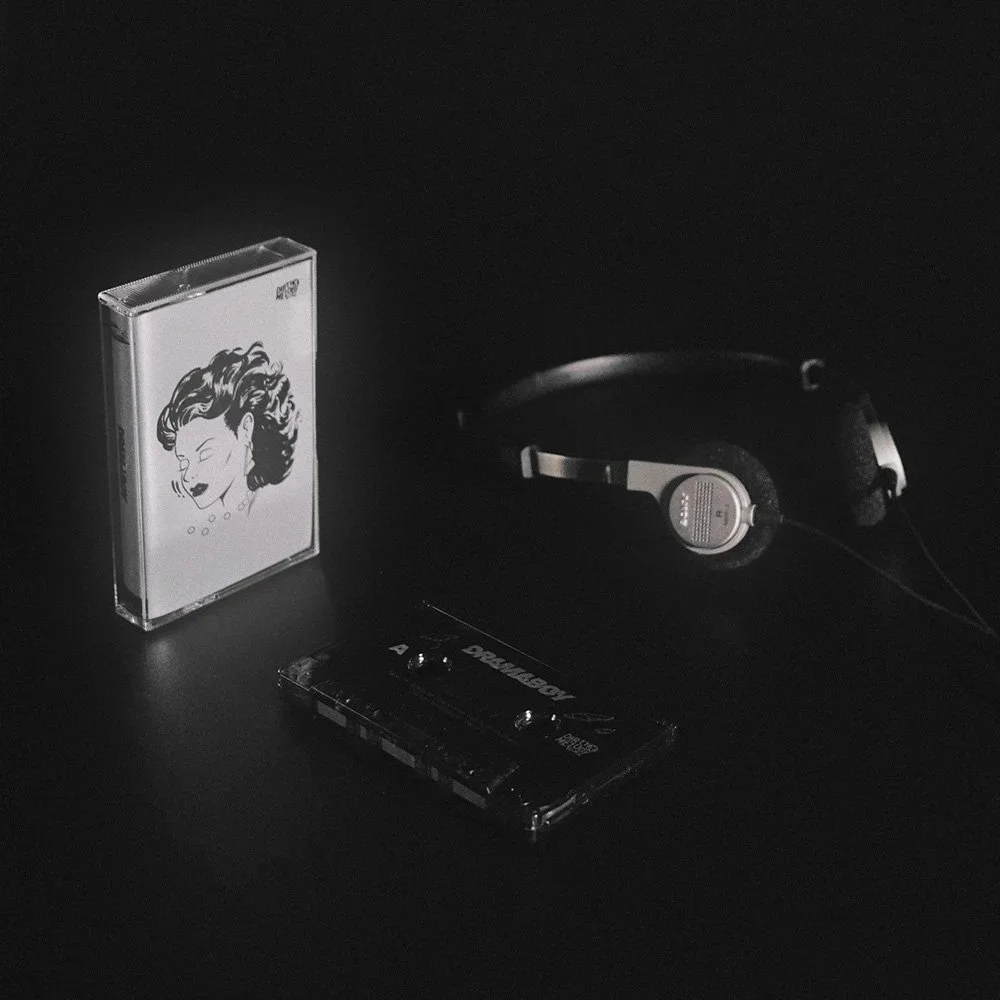 A cassette tape, a pair of headphones, and a cassette case with a woman's face illustrated on it, all placed on a black surface in a dark setting.