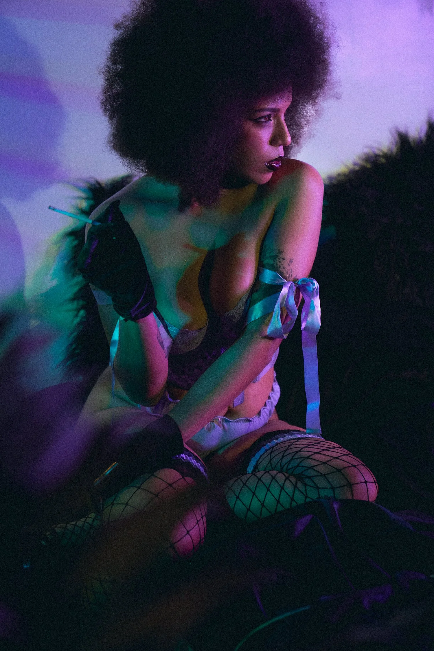 A woman with curly hair, wearing fishnet stockings, satin gloves, and lingerie, sits in a dimly lit room with colorful neon lighting, holding a cigarette.