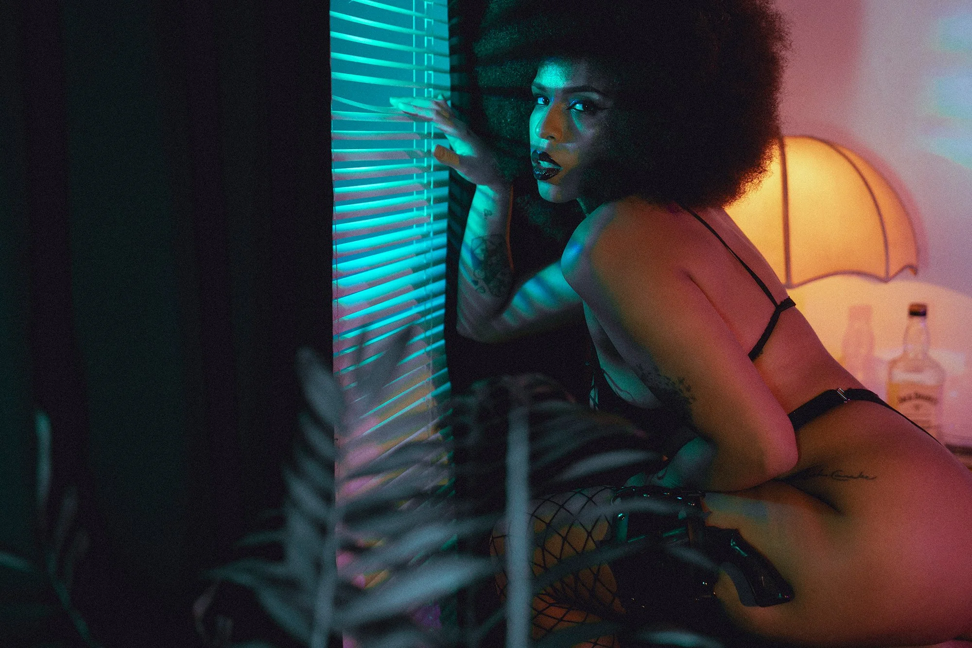 A woman with curly hair and dark makeup looking at the camera, sitting on the floor next to a window with blinds, in a dimly lit room with neon lighting and a lamp in the background.
