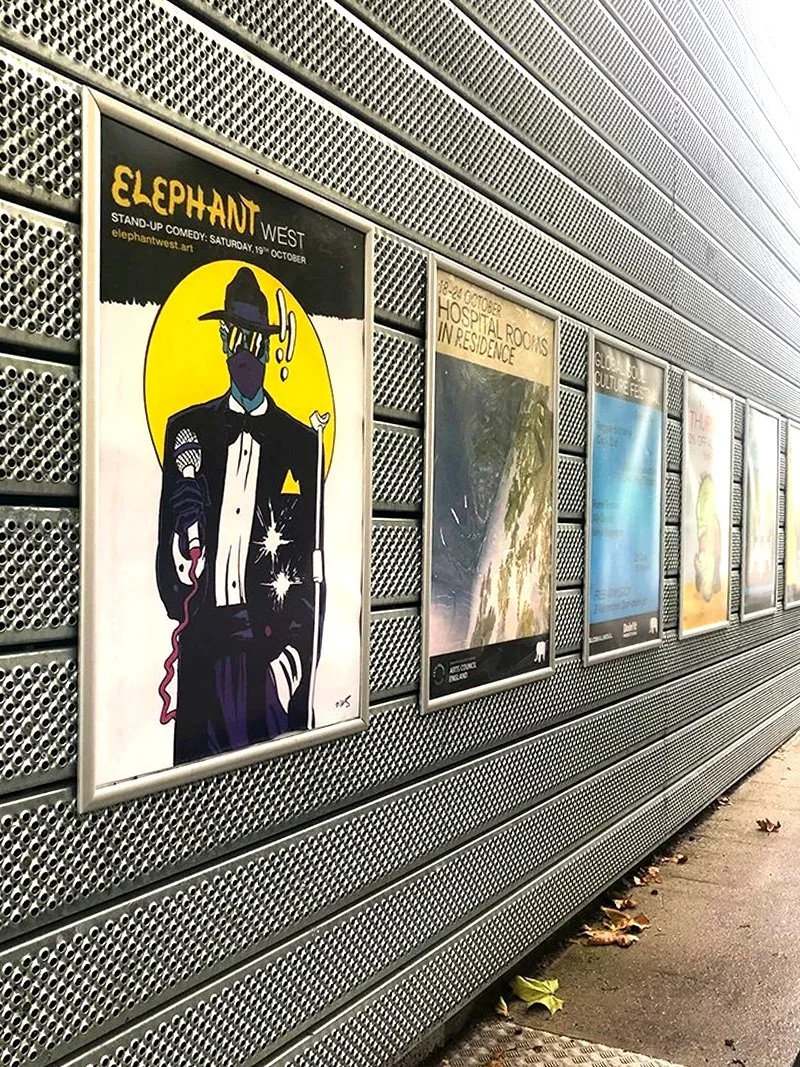 A row of posters displayed on a metallic, perforated wall in an indoor or underground passageway. The first poster promotes Elephant West's stand-up comedy event with a graphic of a stylized figure in a tuxedo and witch hat, holding a cane.