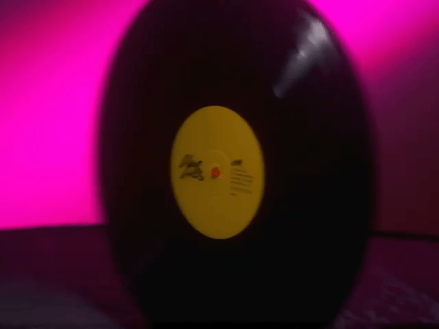 A black vinyl record with a yellow center label, placed against a pink background.