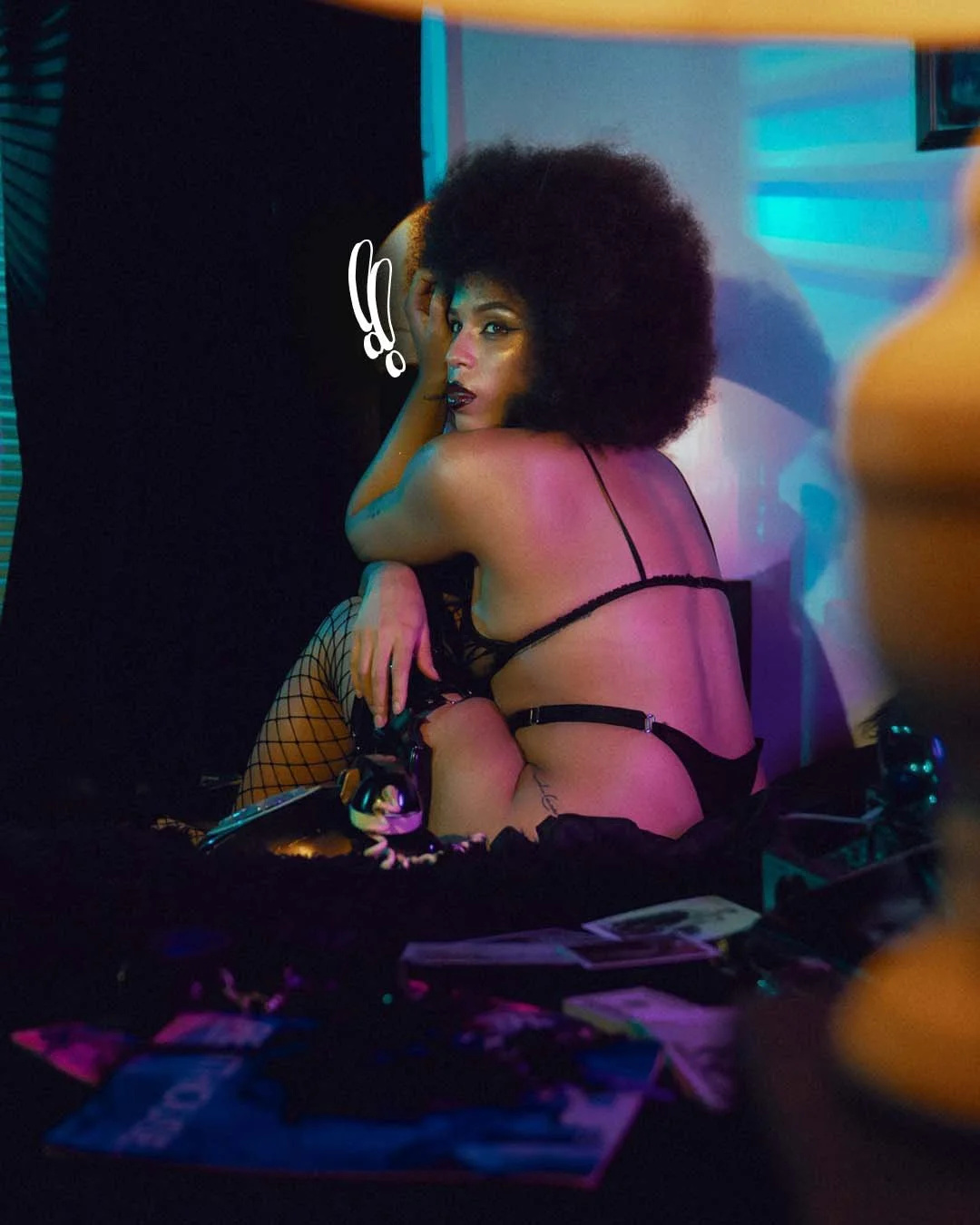 A woman with dark curly hair and dark lipstick, wearing black lingerie and fishnet stockings, sits on the floor in a dimly lit room with colorful neon lighting, surrounded by magazines or photographs.