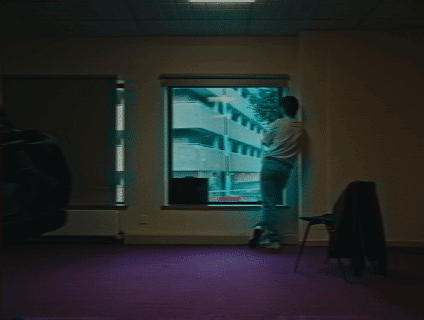 Person standing near a window in a room with purple carpet, empty chair, and a dark object on the left.