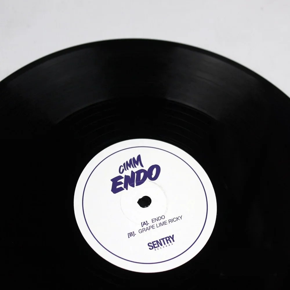 Close-up of a vinyl record with a white label. The label features the text 'CIMMI ENDO' in bold purple font, with song titles 'ENDO' and 'GRAPE LIME RICKY' listed below. The record is black with visible grooves.