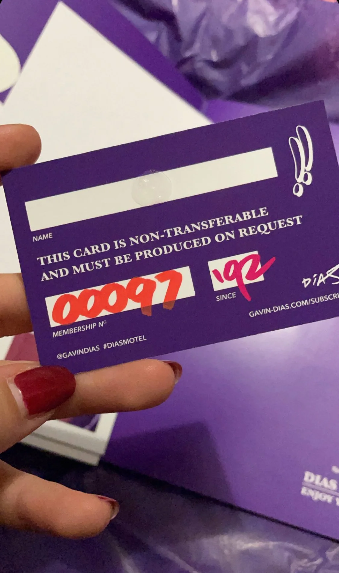 Close-up of a purple membership card with handwritten red and pink numbers, held by a person with red nail polish, purple background, and a plastic bag in the background.
