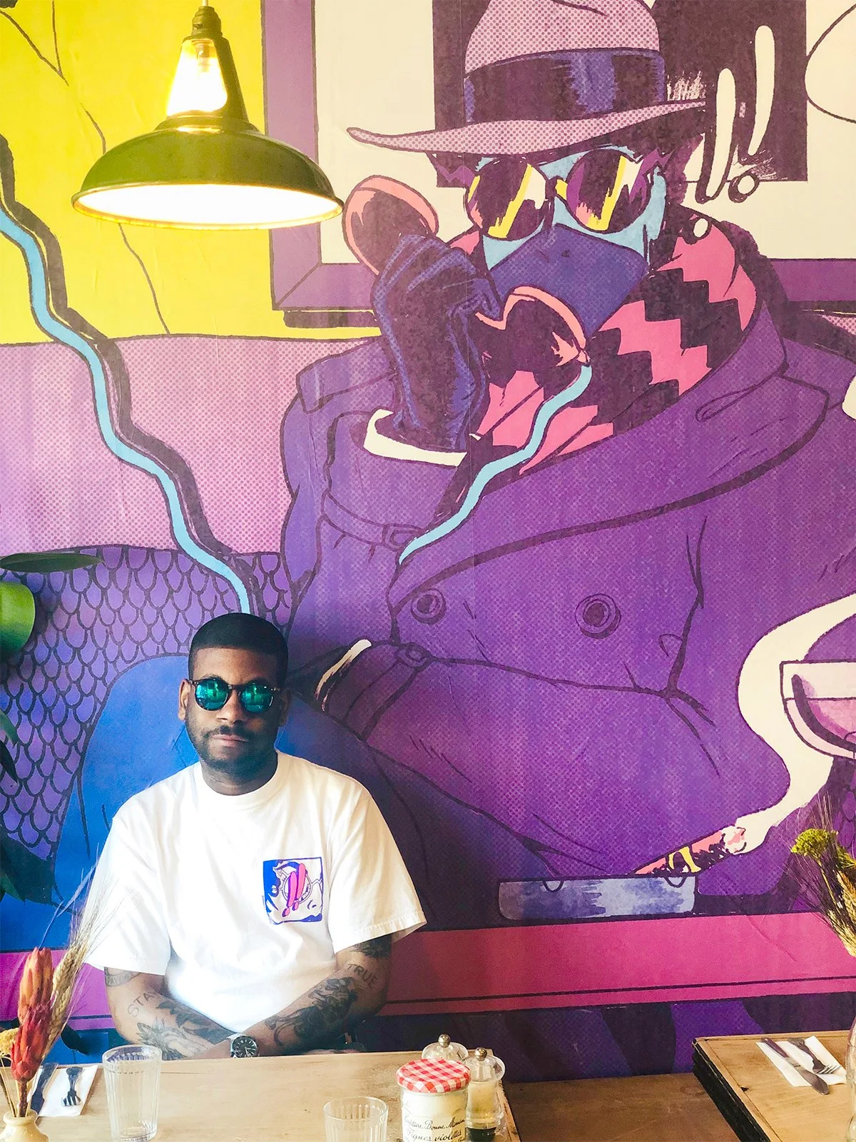 A man wearing sunglasses and a white T-shirt with a graphic, sitting at a restaurant table in front of a colorful mural depicting a stylized woman wearing sunglasses, a hat, and a scarf.