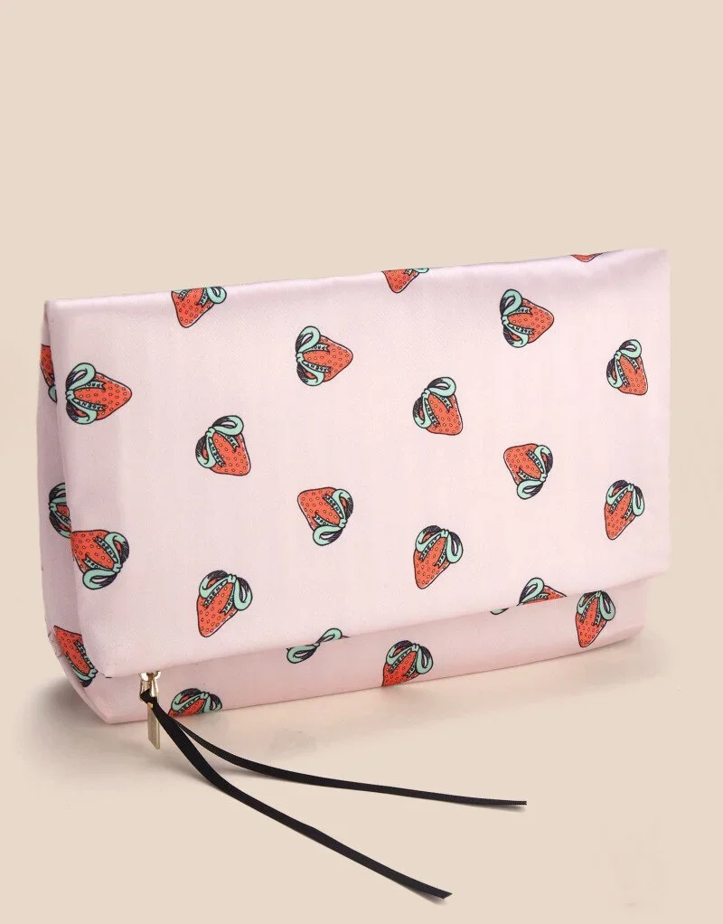 Pink clutch purse with strawberry pattern and black wrist strap on a beige background.