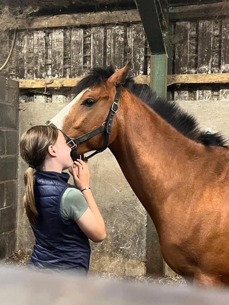 A girl standing face-to-face with a brown horse inside a stable, gently touching the horse's face.