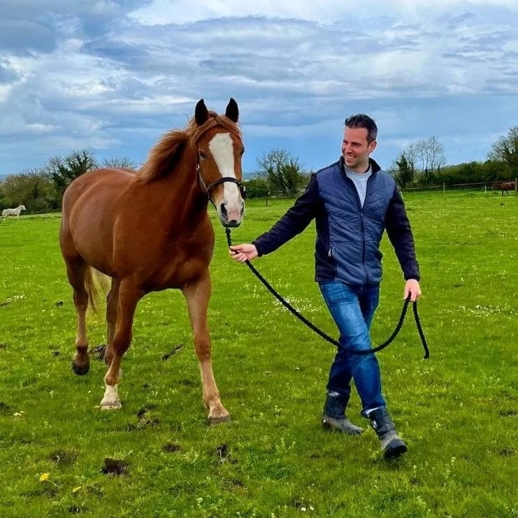 A man in a blue jacket and jeans walking a brown and white horse on a green field under a cloudy sky.