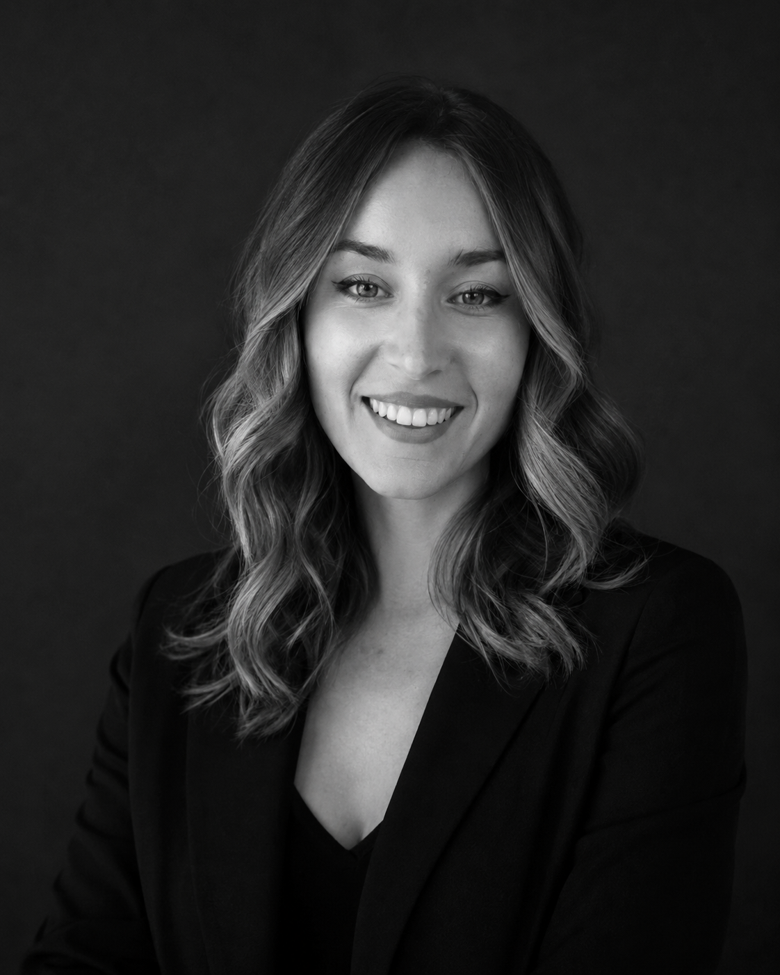 A smiling woman with wavy hair wearing a dark blazer, posed against a dark background in black and white.