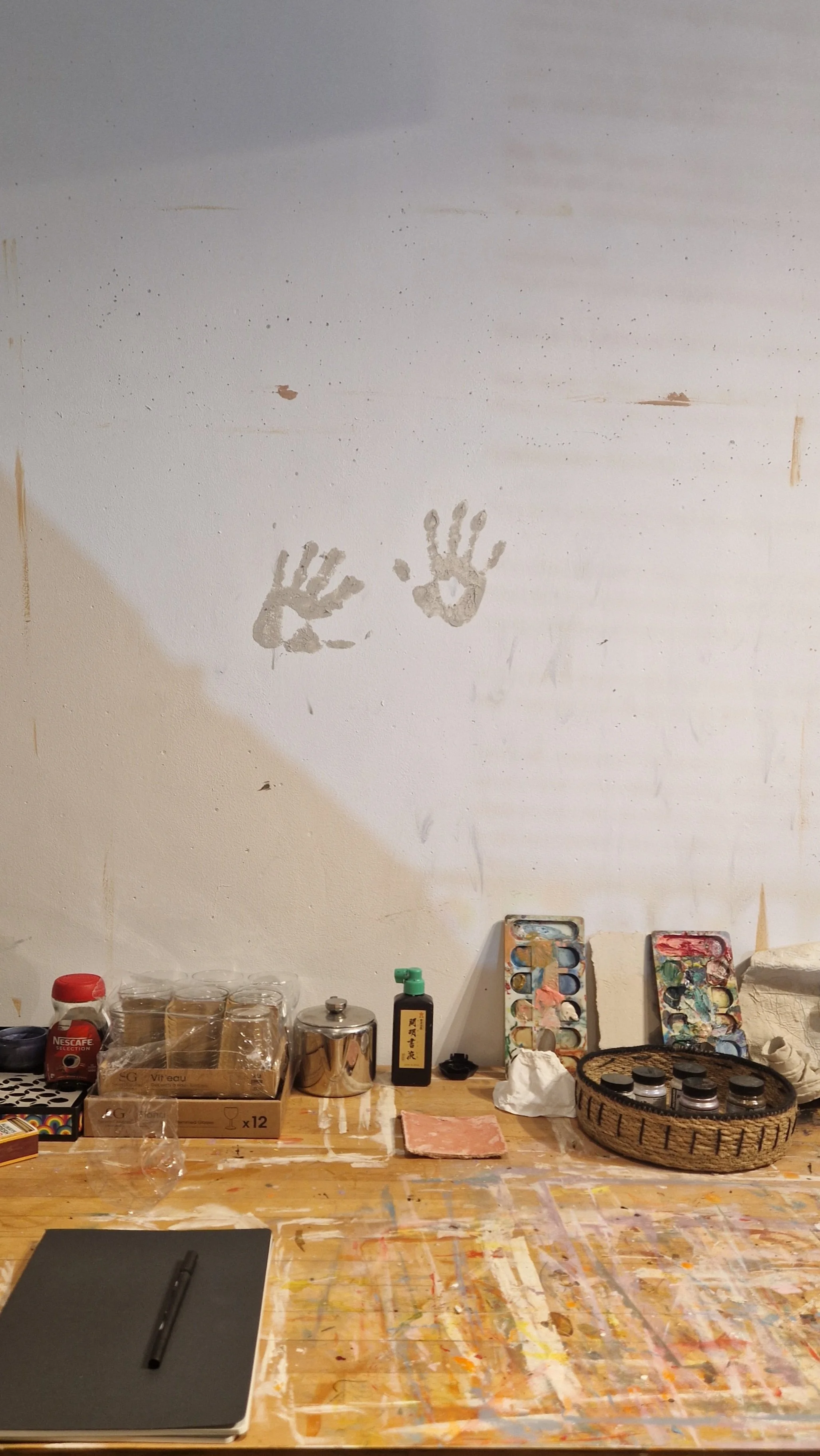 A white wall with two handprints and some small paint splatters. The lower part of the image shows a work table with various art supplies, including paint palettes, jars, a notebook with a pen, and other art tools.