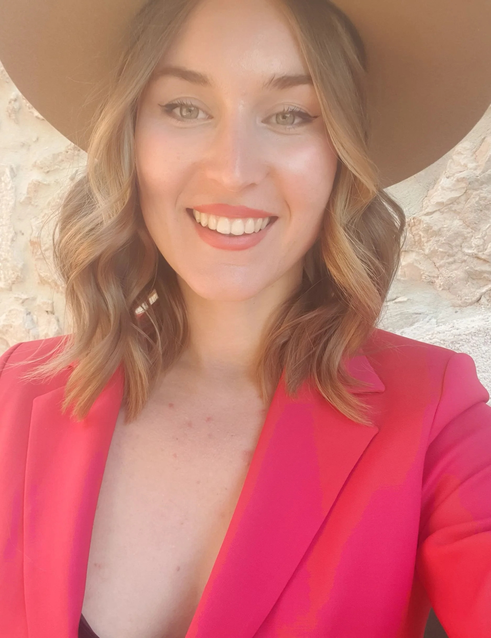 A woman in a pink blazer and large brown hat smiling, with visible cleavage and a textured wall background.