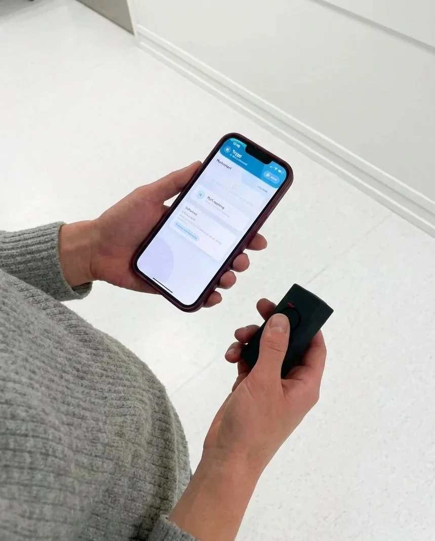 En person holder en smarttelefon i den ene hånden og en fjernkontroll i den andre. Det ser ut til å være en app på telefonen som viser innstillinger for hjemmeteknologi.