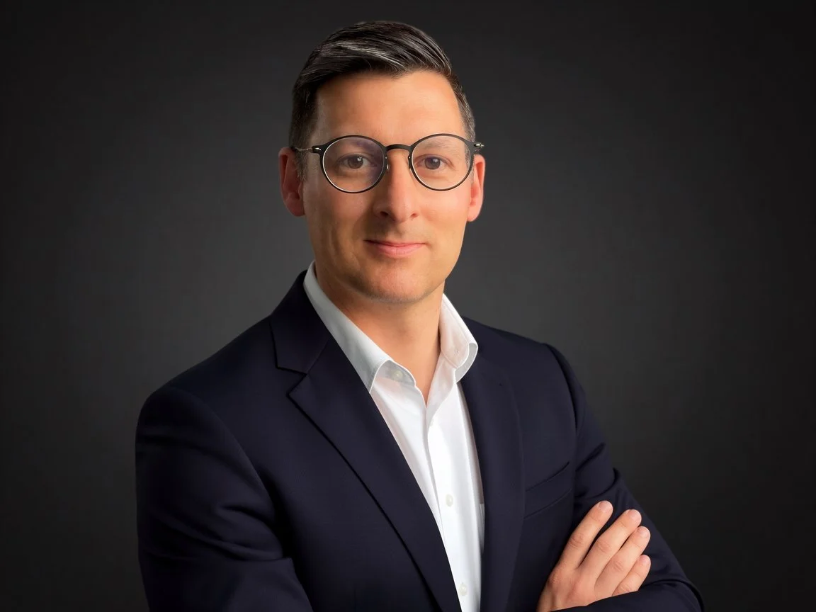 A professional man with glasses, dressed in a dark suit and white shirt, standing with arms crossed against a dark background.