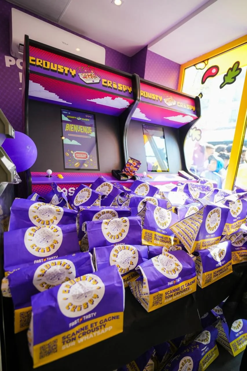 Jeu d'arcade avec sacs de popcorn violet et jaune dans une salle decorated avec des couleurs vives et un ordinateur verte. La signalisation indique 'PEROUSTY CRUSTY' et 'BIENVENUE'.