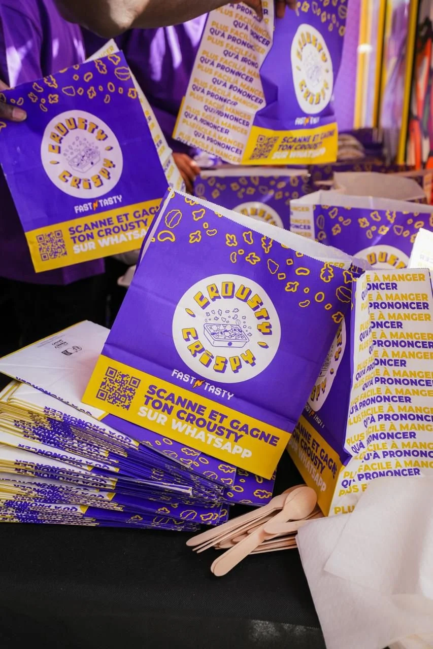 Paquets de croustilles de croustilles avec un design violet et jaune, portant une étiquette demandant de scanner le code QR pour gagner et commandé via WhatsApp.