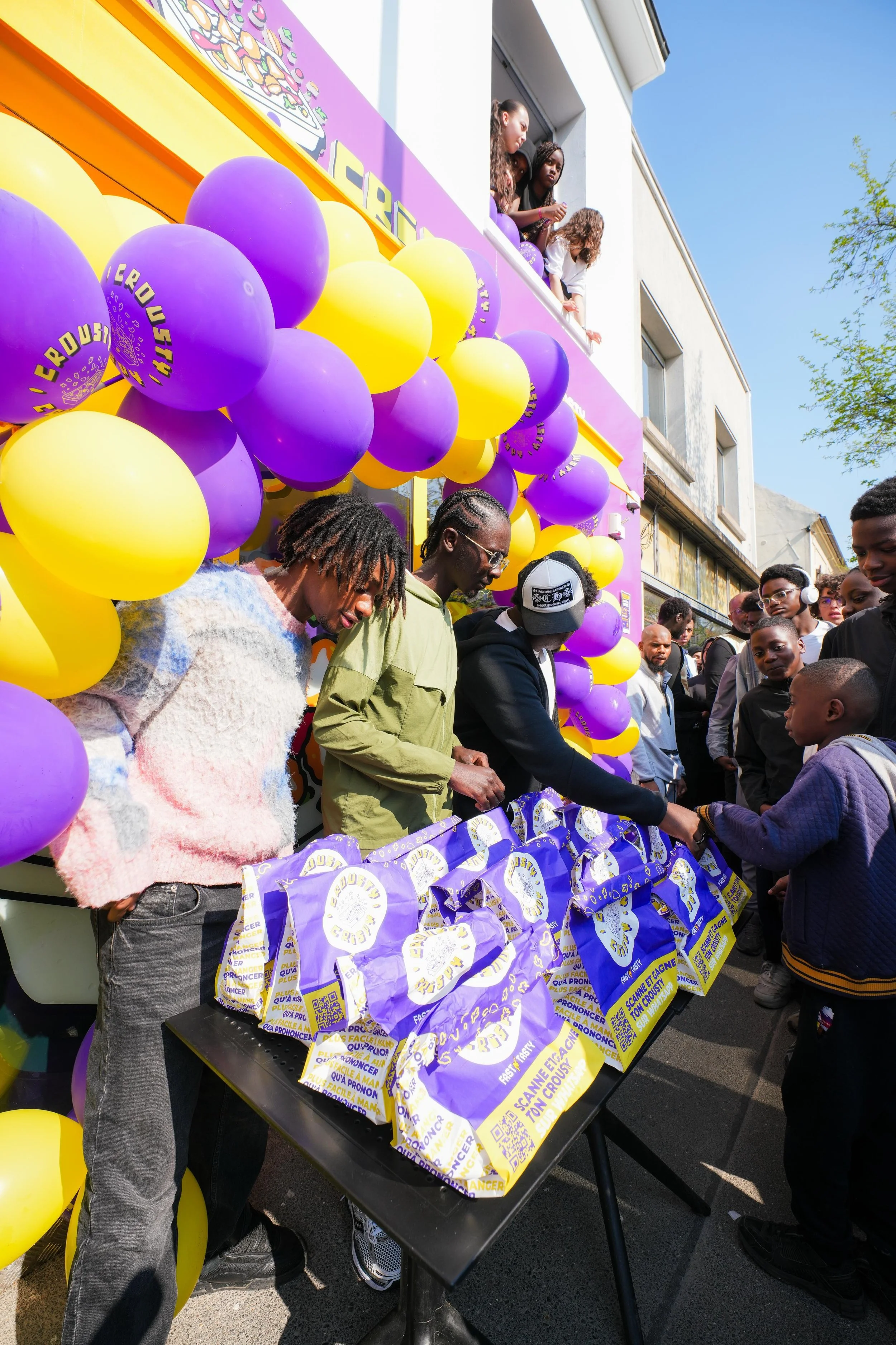 Groupe de personnes distribuant des sacs en plastique violet et jaune avec des ballons assortis à une fête ou un événement en plein air.