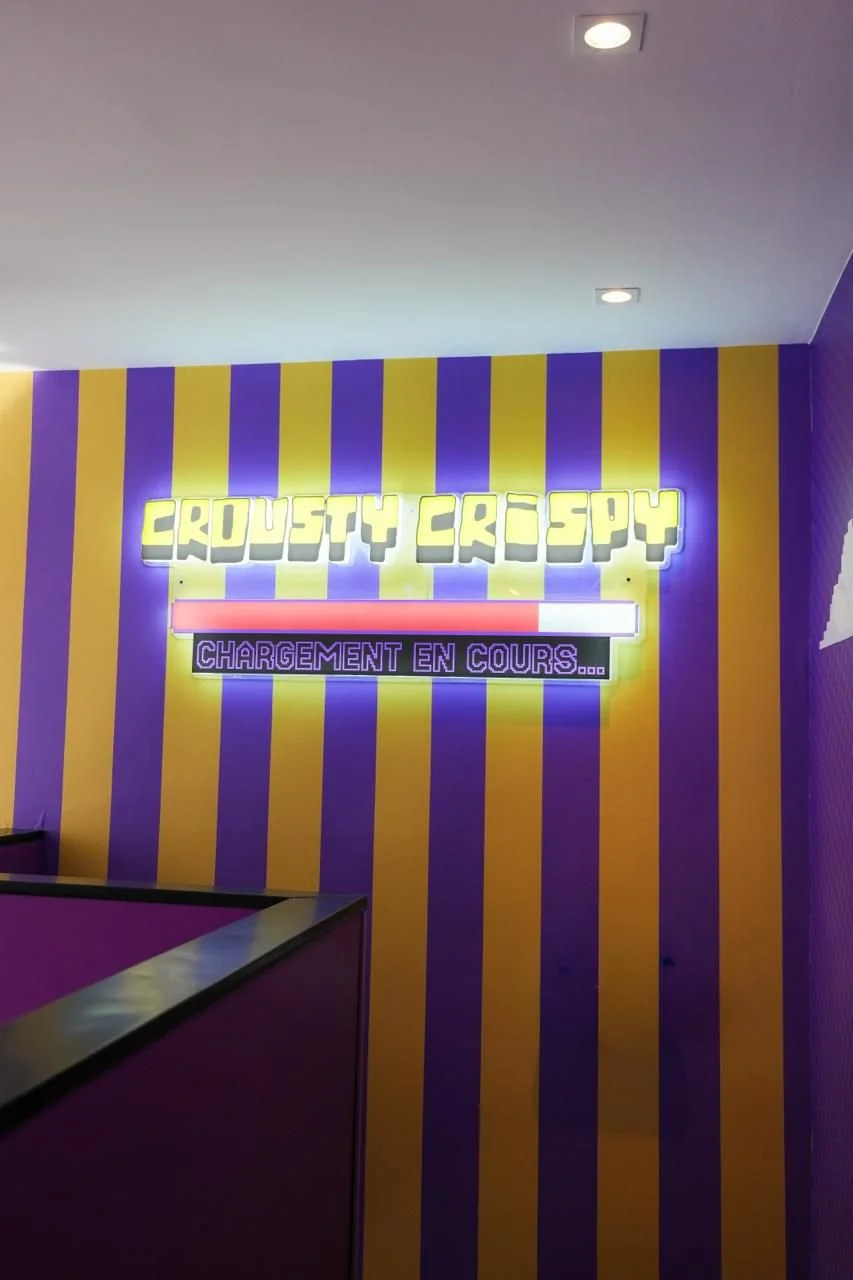 Un mur violet avec des rayures jaunes, une enseigne lumineuse en haut avec le texte "CROUSTY CREEPSY" et une barre de chargement en dessous, accompagnée du message "CHARGEMENT EN COURS..." en lettres violettes