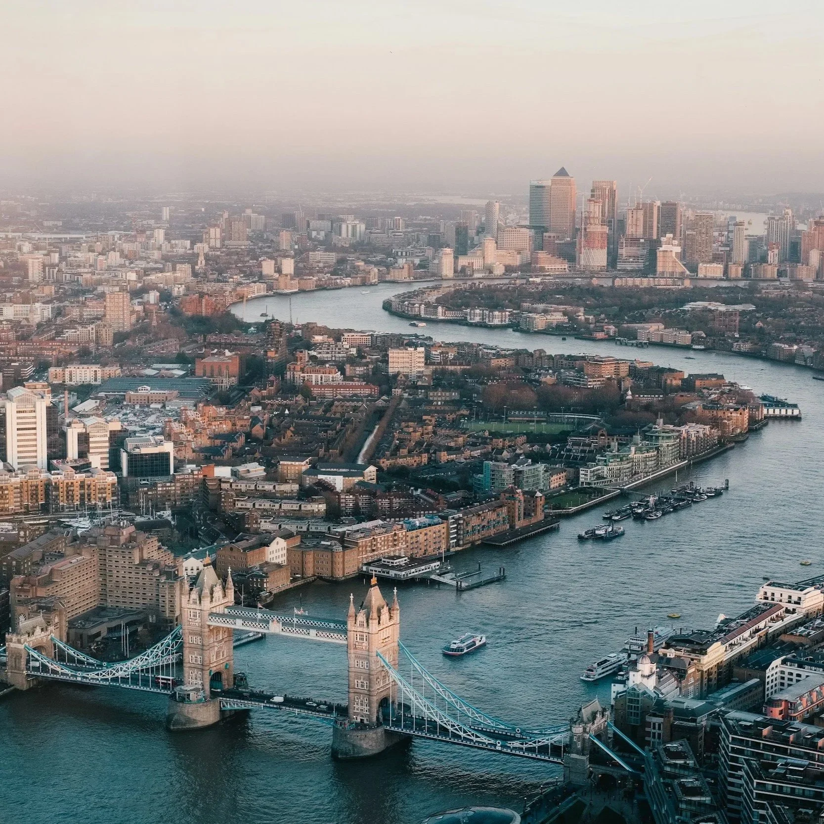 Aerial view of London featuring Tower Bridge crossing the River Thames, with the skyline of skyscrapers and city buildings in the background during daytime.
