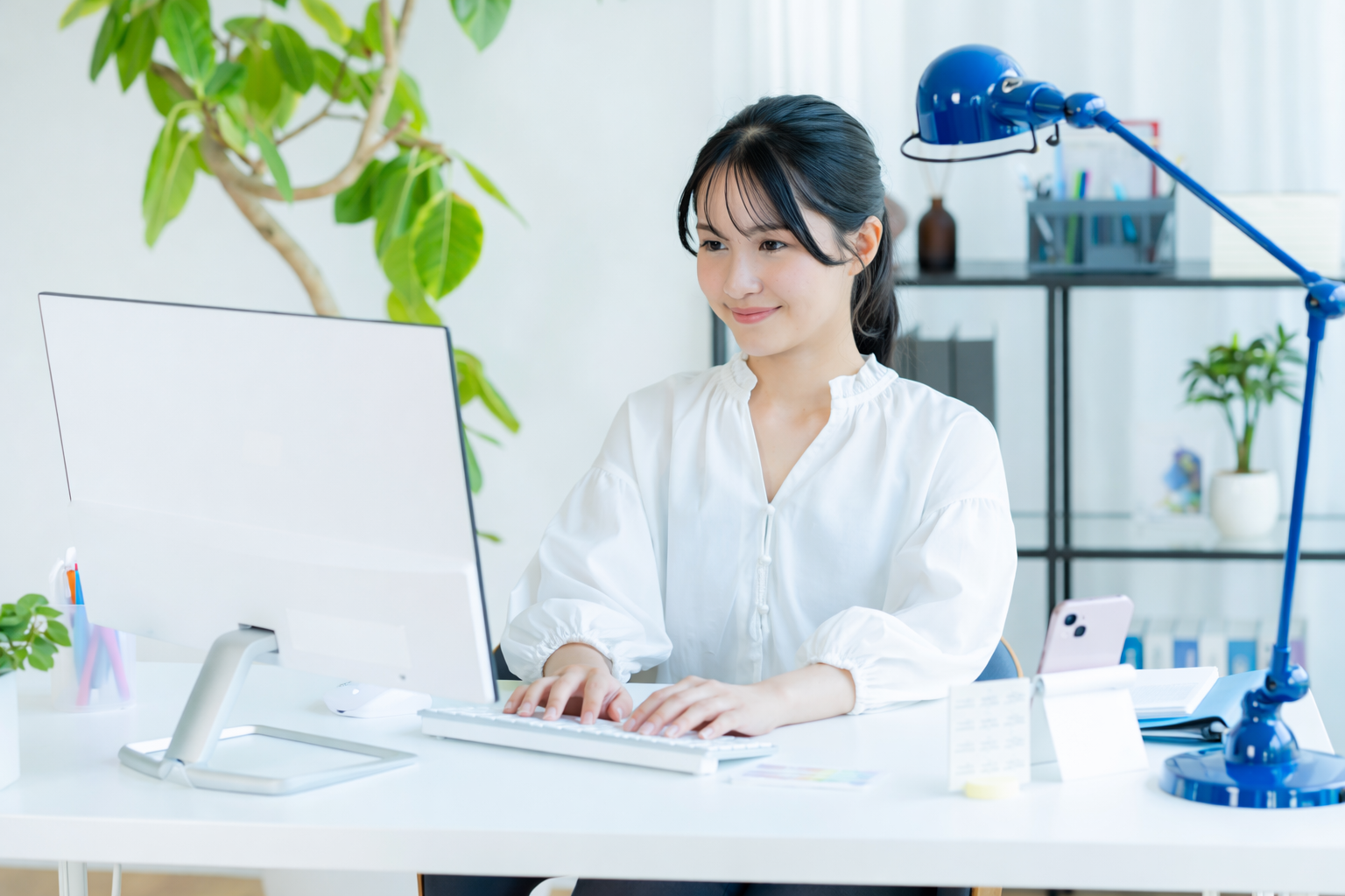 オフィスで仕事をする女性の様子。白いシャツを着用し、パソコンとタイピングしています。背景には植物や書棚が見えます。