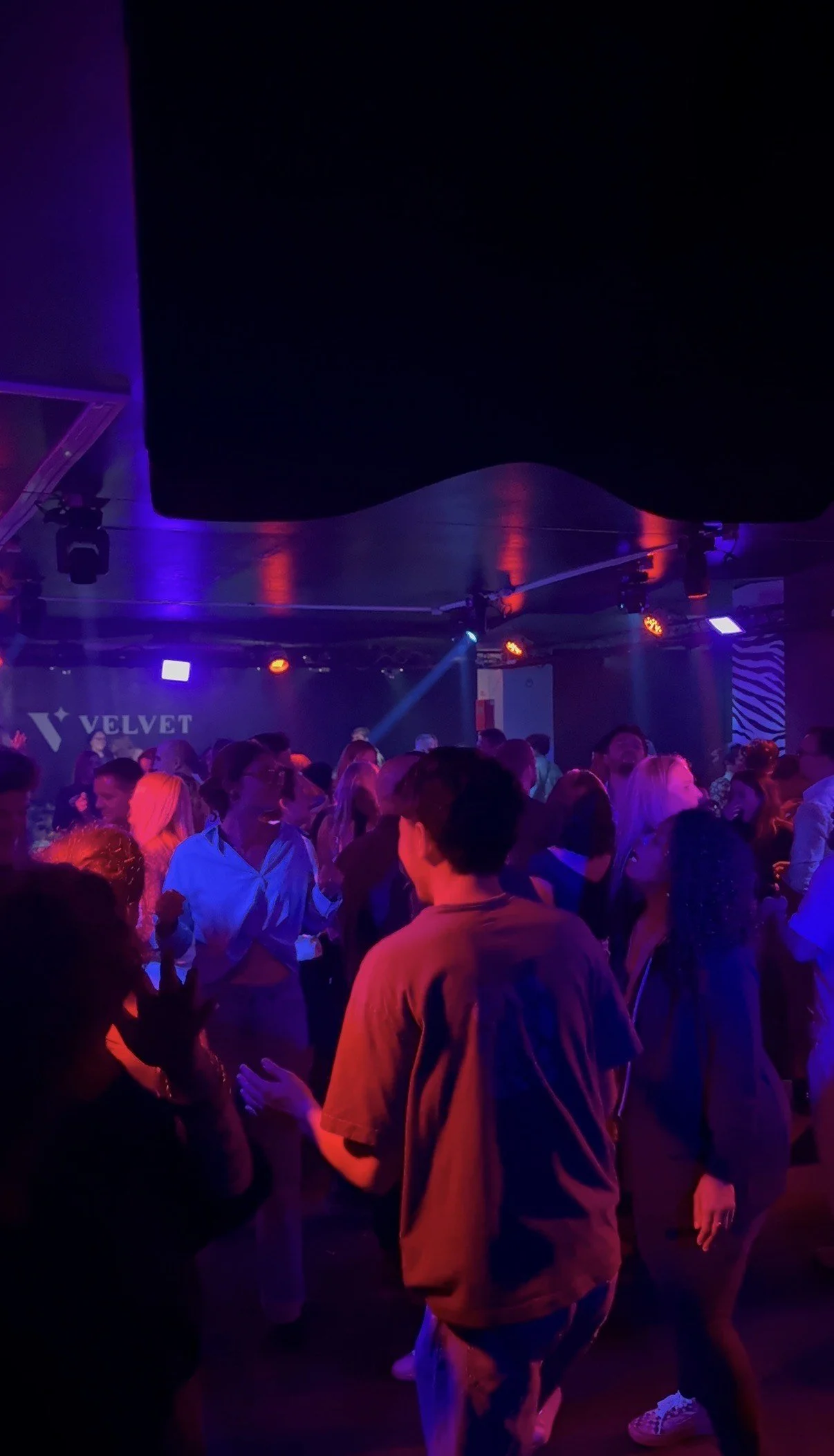 Une scène dans une boîte de nuit avec des gens qui dansent sous un éclairage violet et rouge, avec un mur en arrière-plan portant un logo 'VELVET'.