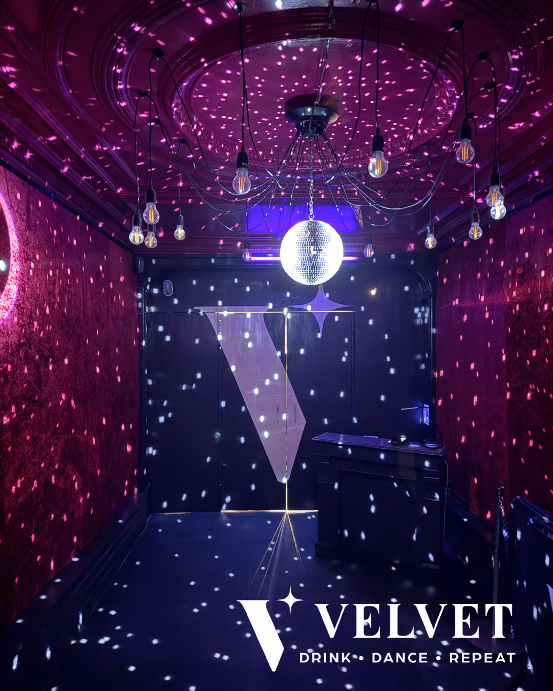 Une salle de danse ou de nightclub avec un éclairage violet, une boule disco, de petites lumières blanches projetant des motifs sur les murs et le plafond, et une bannière avec le logo de Velvet, un lieu de boire, de danser, et de faire la fête.