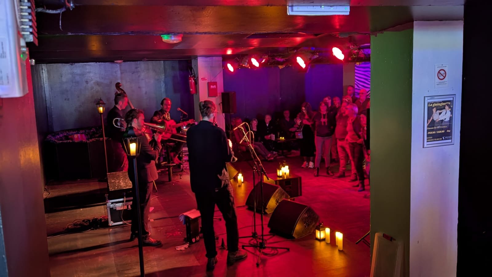 Musiciens jouant en live dans un bar ou un club, avec des spectateurs debout qui regardent la scène, ambiance chaleureuse avec éclairage tamisé et bougies.
