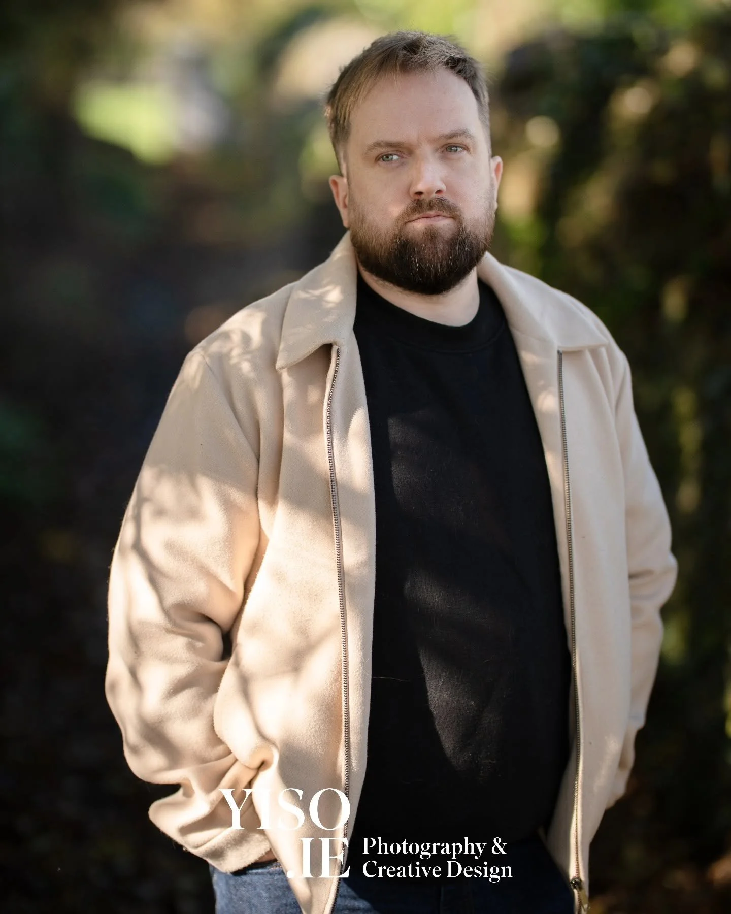 A man with a beard and blue eyes wearing a beige jacket over a black shirt, standing outdoors with blurred trees in the background.