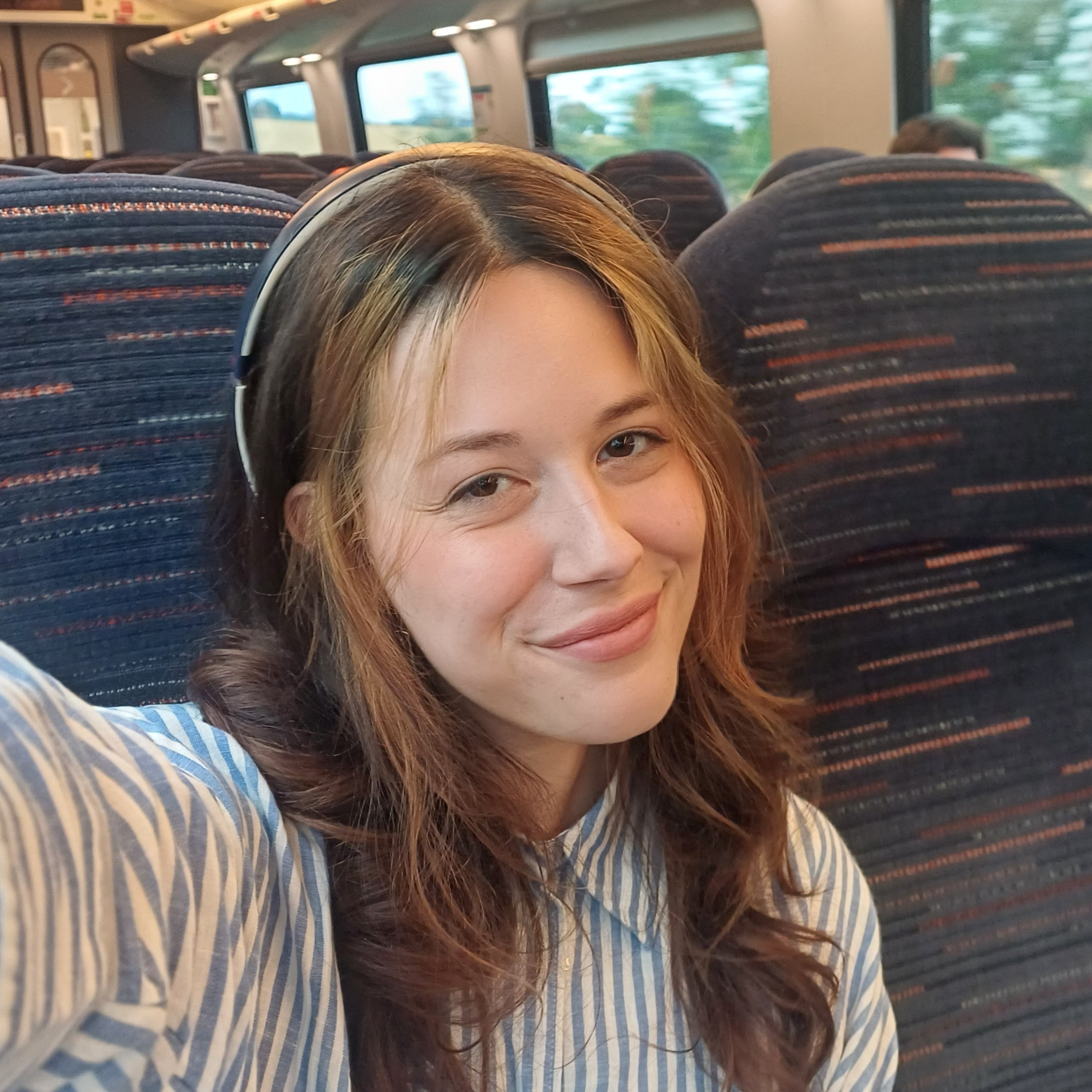 A woman with reddish-brown hair wearing headphones is sitting on a bus. The image captures her face from the nose up, with a background of rows of bus seats and windows showing a blurred outdoor scenery.
