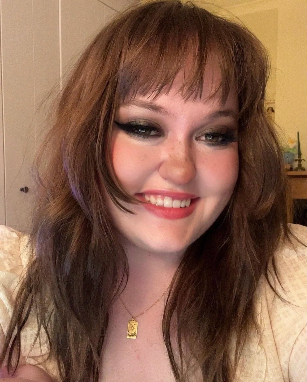 Close-up of a smiling young woman with long, wavy brown hair and makeup, wearing a gold necklace with a rectangular pendant, in a room with beige and yellow walls.