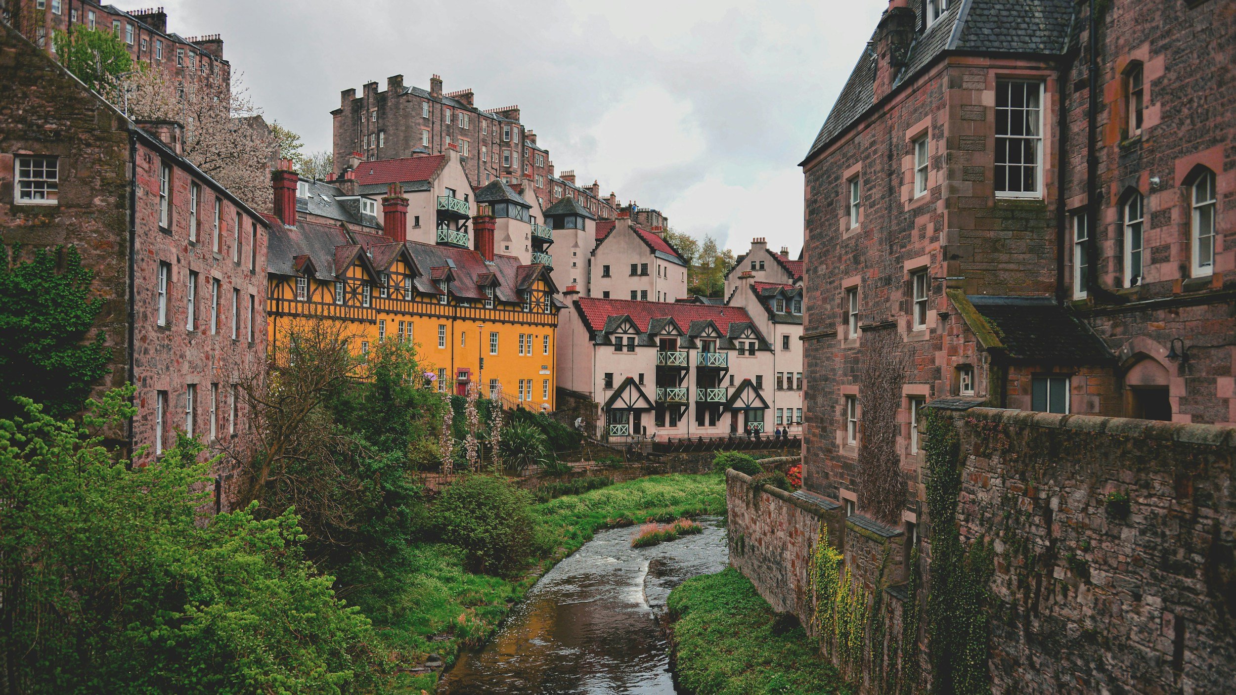 Historic European-style buildings with colorful facades and stone walls along a small river with greenery.