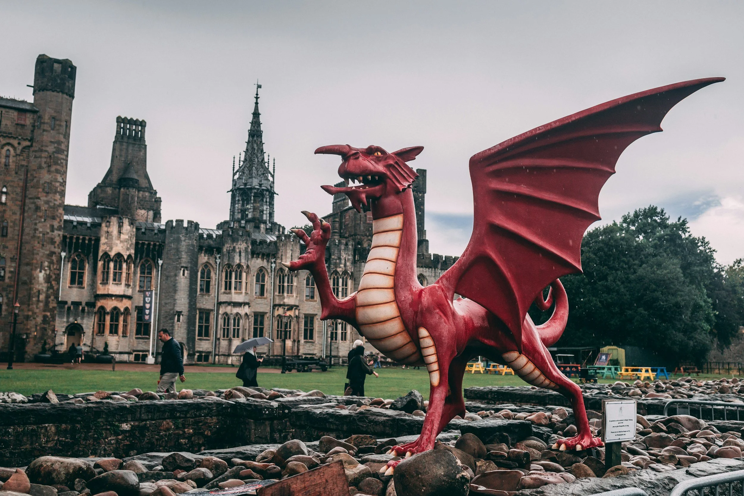 A red dragon statue with large wings and a menacing expression stands in a park in front of a historic castle-like building. People are walking nearby, some holding umbrellas, on a cloudy day.