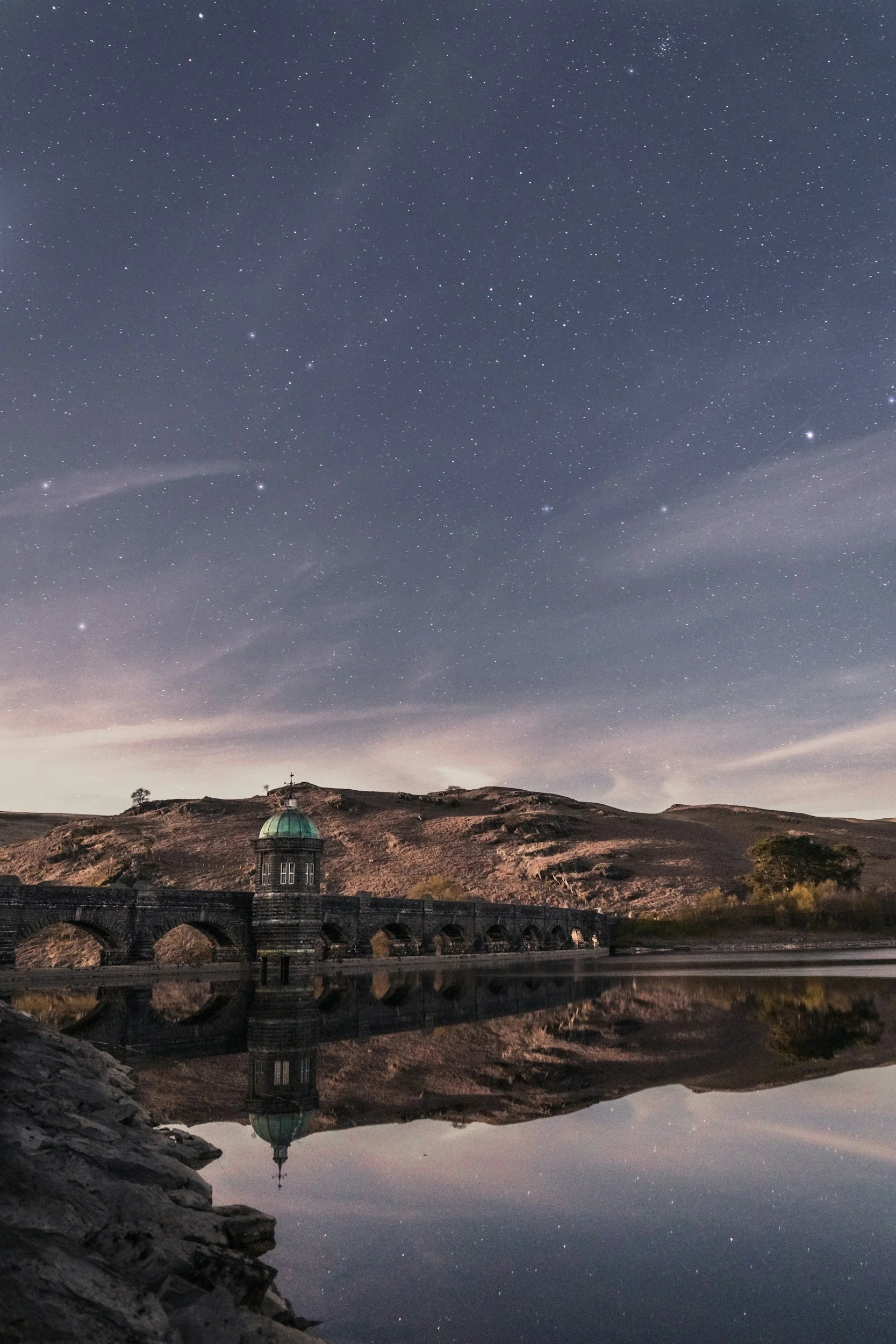 Nighttime landscape featuring a starry sky over a bridge and a historic building with a domed roof, reflected in a calm body of water.